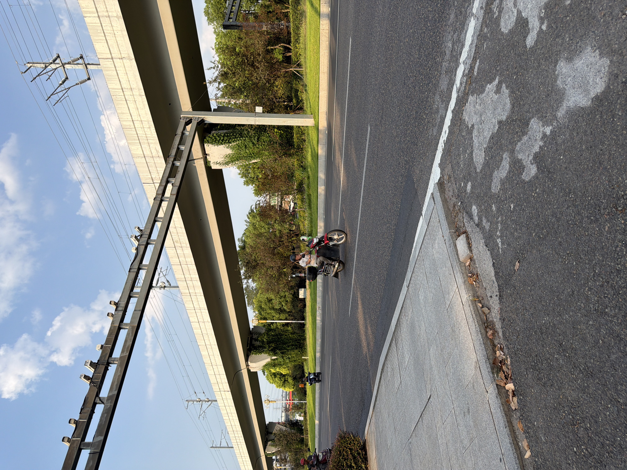 In Jiangjin, China, under a clear blue sky with scattered clouds, an overpass for an elevated railway or highway dominates the background. The overpass is a massive concrete structure supported by substantial pillars, with power lines and what appear to be security cameras visible along its top edge. Below the overpass, a person is riding a red motorcycle in the right lane of a paved road. The motorcycle has a black rear storage box. Further in the distance, two other motorcycles are visible on the same road, one in the left lane and one further back in the right lane. The road is bordered by a sidewalk made of gray paving stones on the left, with some dried leaves and debris along the edge. Beyond the road and sidewalk, there is a grassy median with trees and bushes. The overall scene depicts urban infrastructure and everyday transportation.