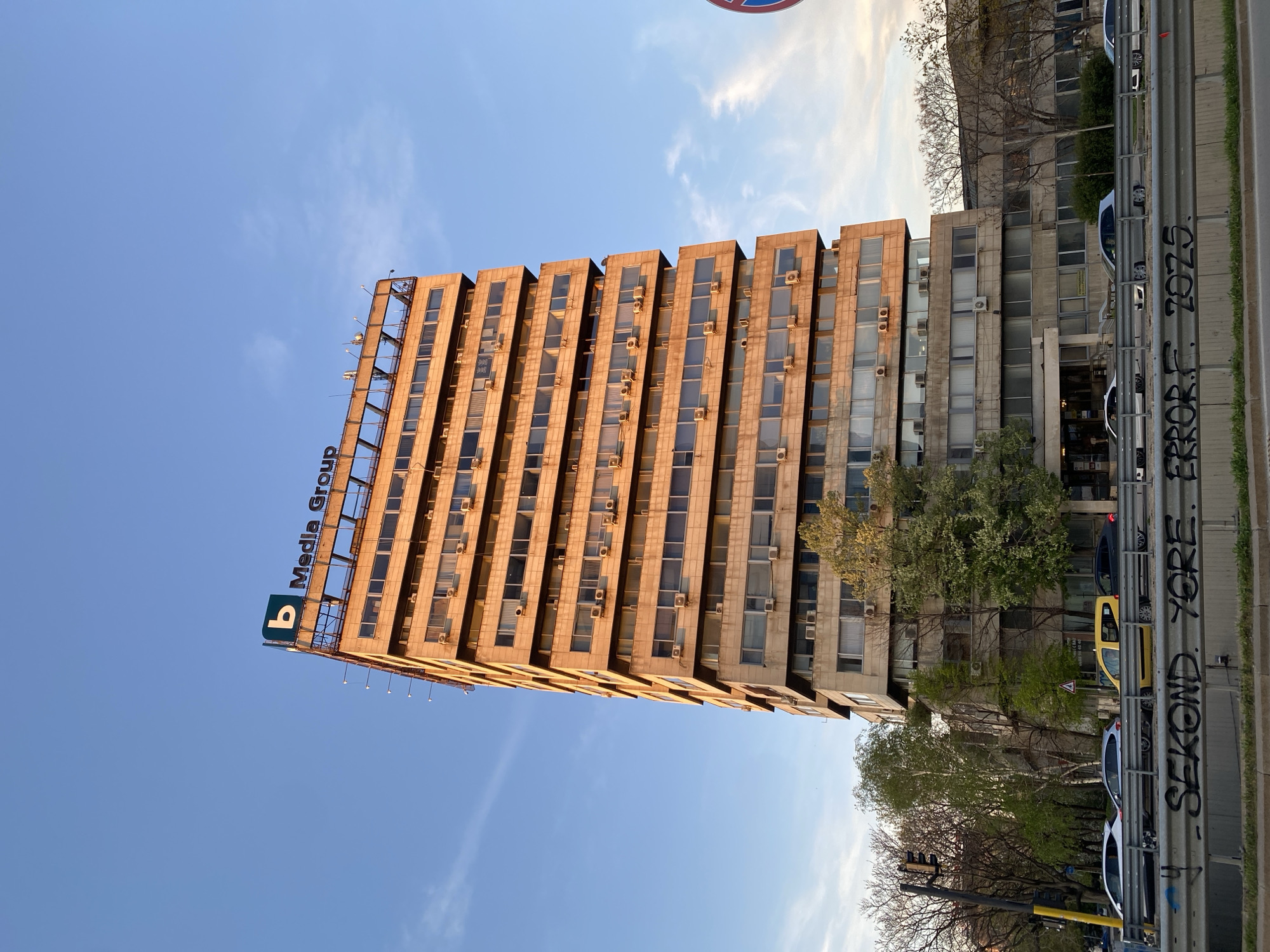 The image shows the B Media Group building in Sofia, Bulgaria. The building is a tall, rectangular structure with a grid-like facade of windows and vertical concrete dividers. Many of the windows have individual air conditioning units attached. A sign reading "B Media Group" is visible on the top of the building. To the right, an older, shorter building is visible. A road runs along the right side of the image, with a metal guardrail covered in graffiti. Several parked cars and trees are visible in the foreground. The sky is blue with scattered clouds.