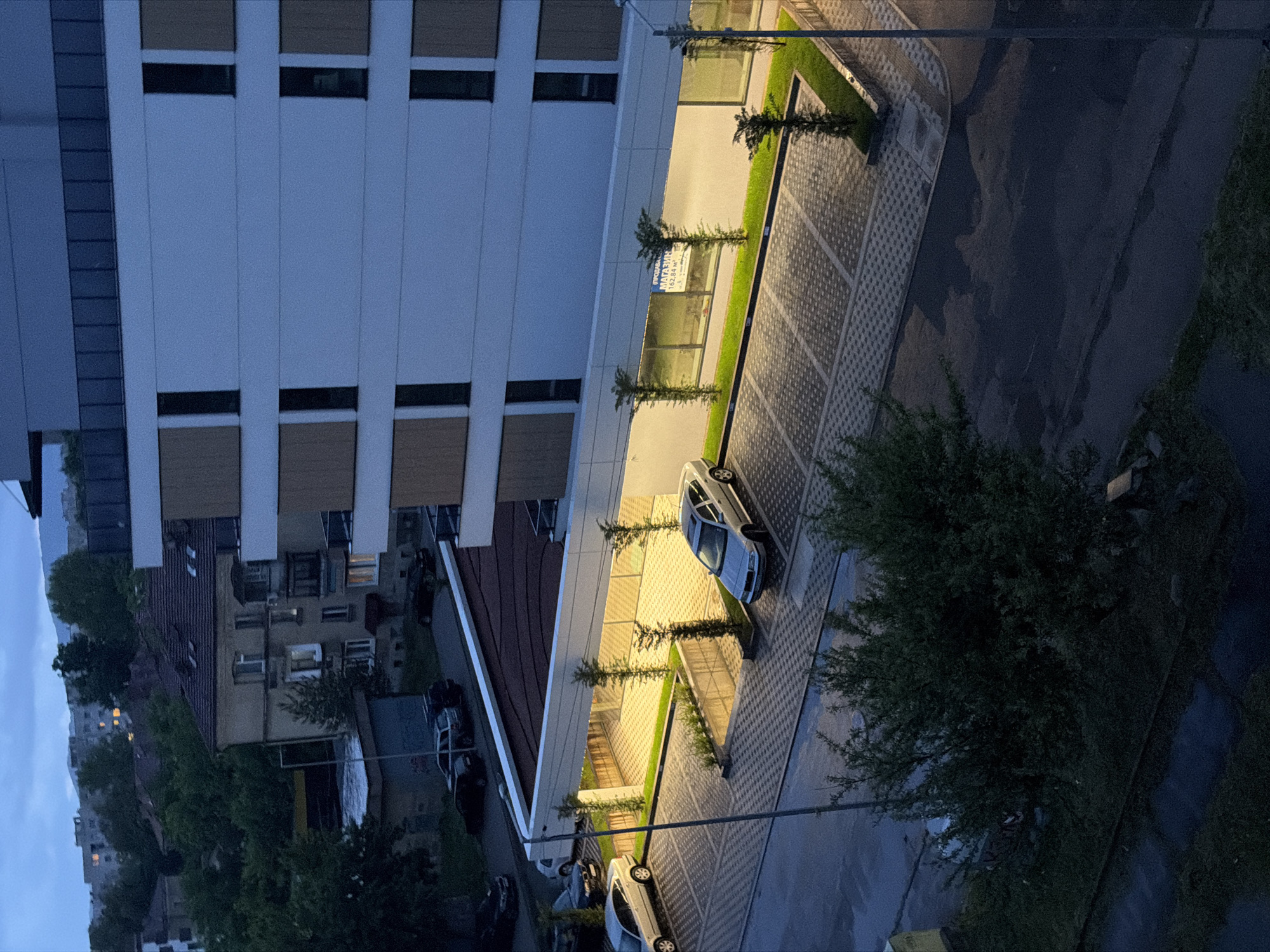 This image captures an urban scene at dusk or night, viewed from an elevated perspective looking down. The scene is dominated by a modern, multi-story building on the left, featuring a facade of light-colored panels interspersed with dark horizontal window sections. Below this building, there's a brightly lit ground-level area with patterned pavement used as a parking lot, illuminated by warm artificial lights. Several cars, including at least two white sedans, are parked here. Small, slender trees line a strip of grass alongside the illuminated pathway.
To the right of the parking area, a darker asphalt road is visible, bordered by dense, dark green foliage in the foreground and further into the background. Beyond the modern building and parking lot, the background reveals older, more traditional residential buildings, some with lights on in their windows, suggesting habitation. In the far distance, under a dim twilight sky, hills or more distant urban structures can be seen.
No people are visible, and no specific event or activity is taking place; the scene is static. The artificial lighting creates a strong contrast with the fading natural light of the evening.
Visible text on a sign in the illuminated area reads "МАГАЗИН" (which translates to "Shop" or "Store"), followed by numbers such as "16244" and "-3".