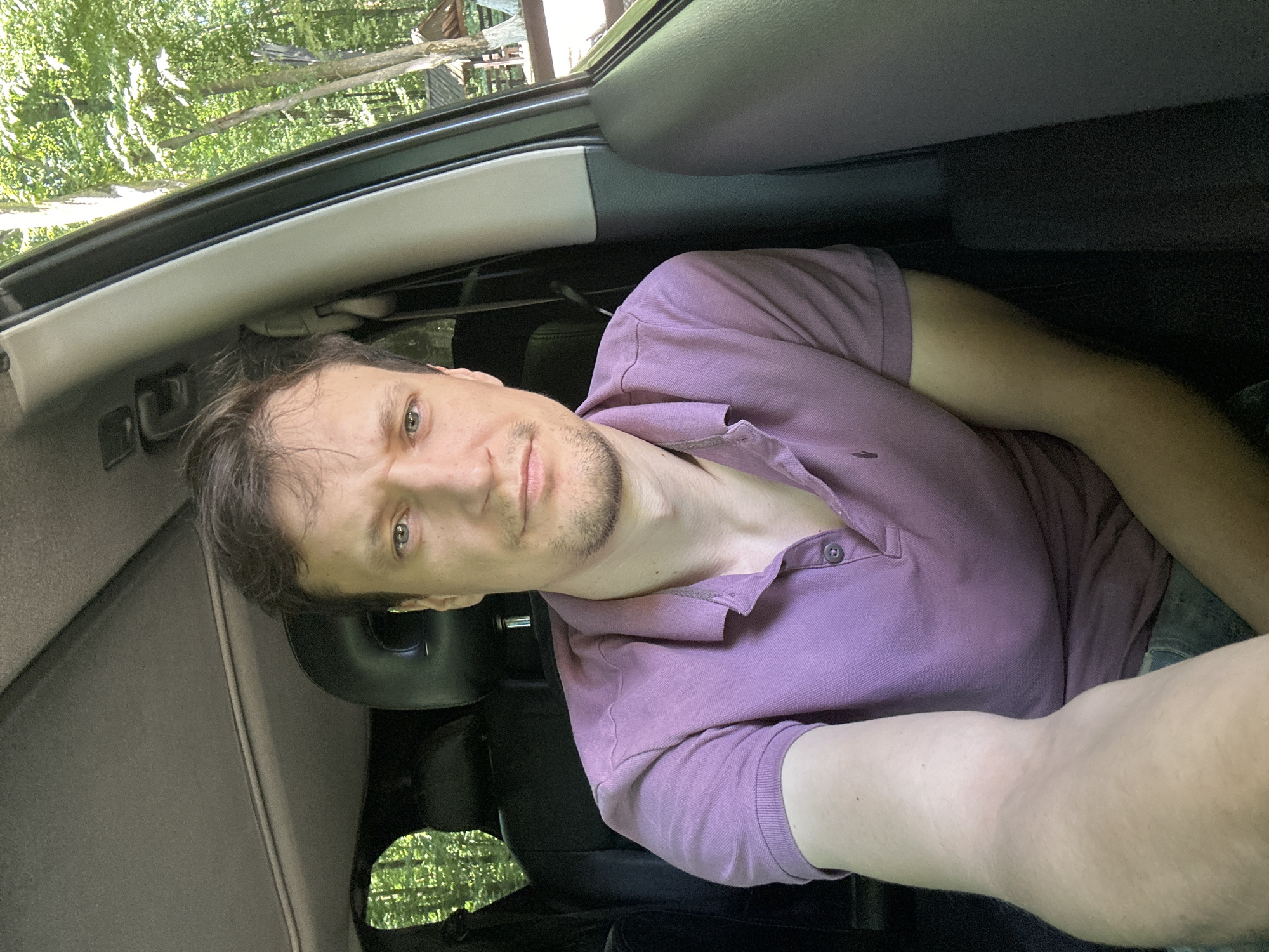 A man with short, dark hair and a light complexion is taking a selfie from the driver's seat of a car. He is wearing a purple polo shirt and appears to be looking at the camera with a neutral expression. The background visible through the car window suggests a natural, wooded environment, with trees and foliage under a bright sky. The interior of the car is dark, with black leather seats visible behind him. The car is likely parked on a road or in a parking area surrounded by trees, possibly in the vicinity of Shatava, Ukraine, given the presence of trees and vegetation characteristic of a rural or park-like setting.