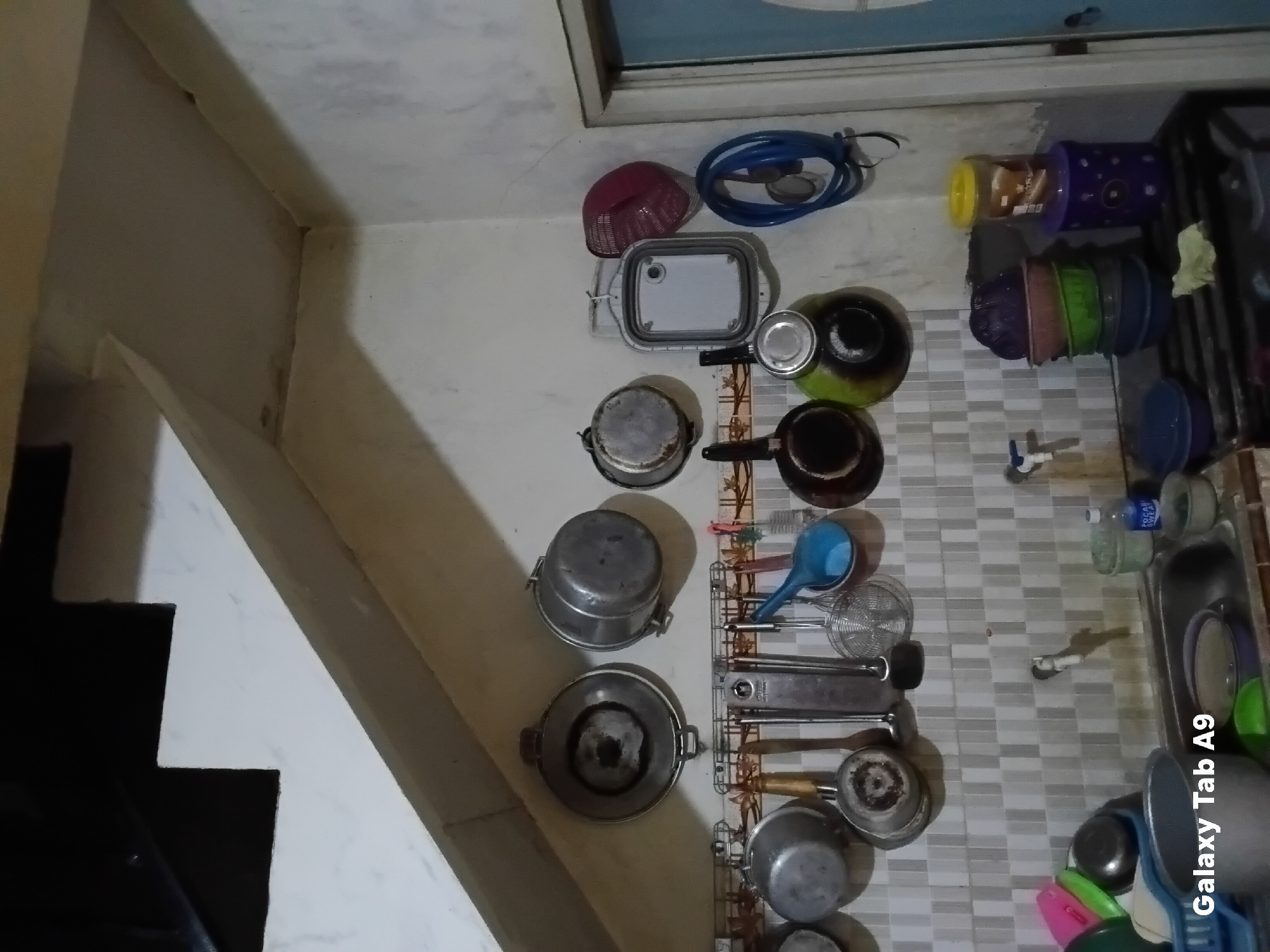 The image depicts a kitchen scene, likely in Trenggalek, Indonesia. The kitchen is sparsely decorated with various cooking utensils hanging on the wall. These include pots, pans, ladles, and strainers. A faucet is visible above a sink, and several containers, possibly for food storage, are stacked on a counter. The overall impression is one of a functional, lived-in kitchen.