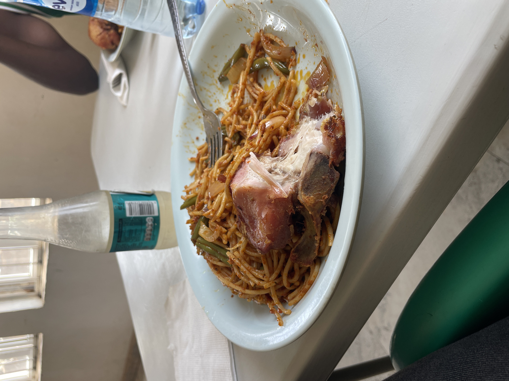 A partially consumed plate of spaghetti with meat sits on a white table. A silver fork rests in the spaghetti. Next to the plate are a bottle of water and a clear glass bottle containing a green liquid. A person is seated at the table wearing black tights and dark jeans. The location is possibly in Jos, Nigeria, based on visual indicators like the food presentation.