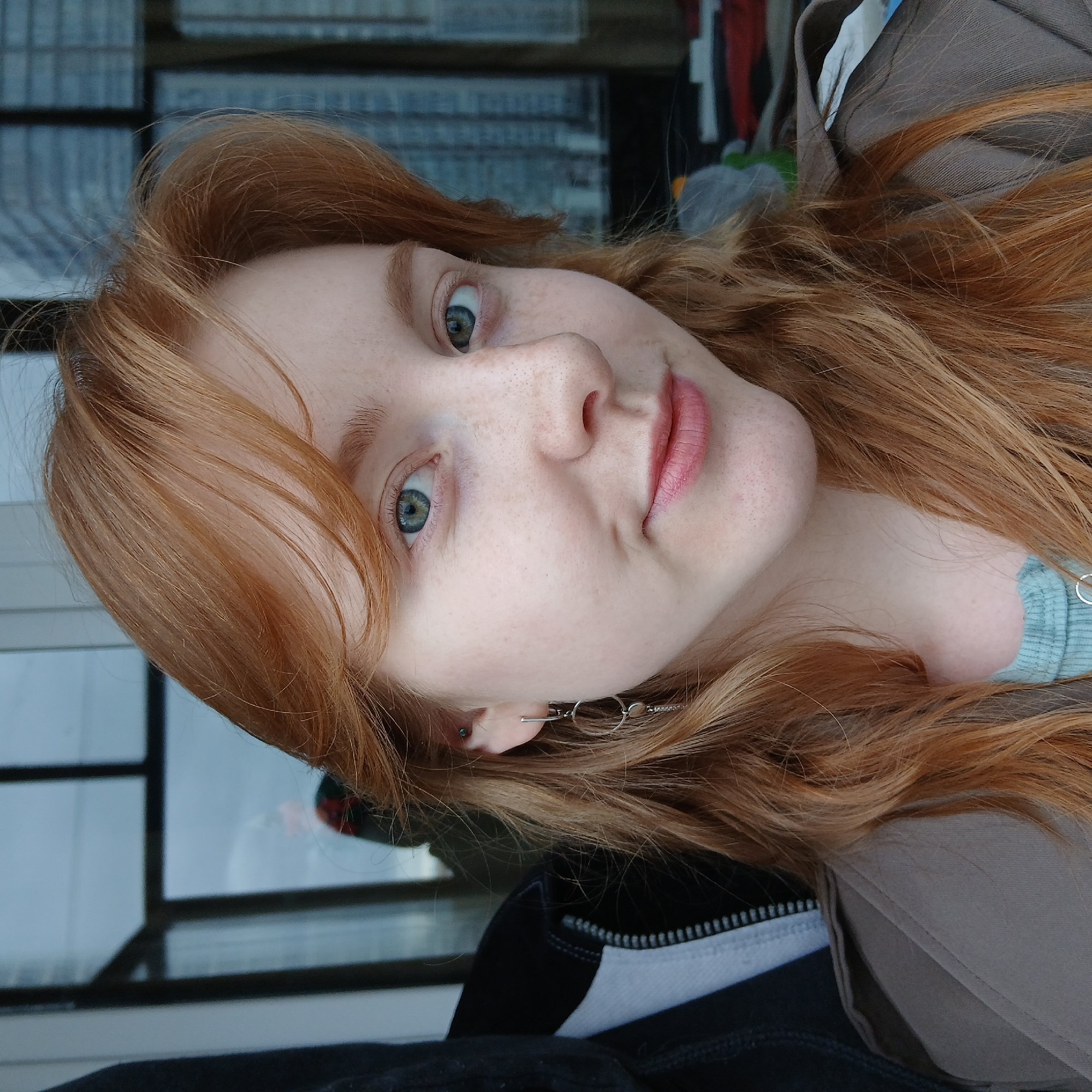 A young, light-skinned woman with long, reddish-blonde hair is positioned in front of a window. She gazes directly at the camera with an expression that could be interpreted as a slight pout or smile. Her face is freckled, and she has light eyes. She wears earrings, and portions of her clothing, including what appears to be a brown jacket over a light blue top, are visible. In the background, a window offers a blurred view, possibly of buildings. The location is most likely an indoor space, and based on the prompt, it is claimed to be in Yekaterinburg, Russia.