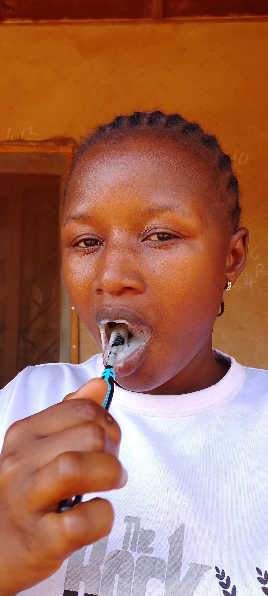 A dark-skinned woman with braided hair is brushing her teeth with a black and blue toothbrush and white toothpaste. She is wearing a white t-shirt with a printed design, possibly the word "The" followed by another word beginning with "R" and decorative laurel branches. She is located near a door frame set against a warm, tan-colored wall with some writing on it. The location is likely Jos, Nigeria, given the prompt's instruction.