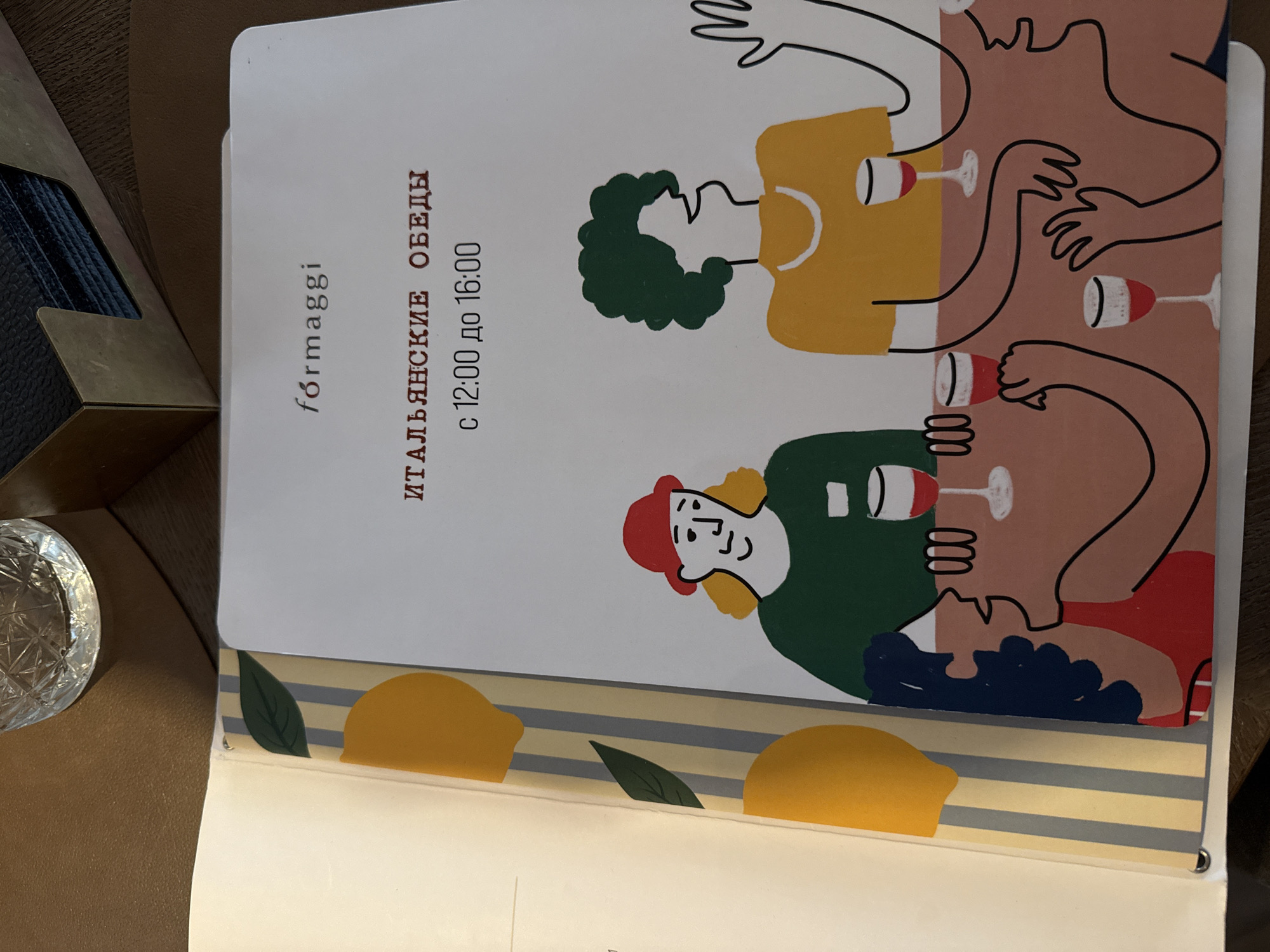 A close-up shot reveals a menu from the restaurant "Formaggi" in Krasnoyarsk, Russia. The menu is open, displaying a cartoon-style illustration of four people around a table, seemingly enjoying wine. The people are depicted with different hair colors and wearing different colored shirts. The menu also includes text in Russian, indicating "Italian lunches" from 12:00 to 16:00. Underneath the menu, a yellow-and-gray striped design with stylized lemons and leaves is visible. A partial view of a textured metallic or composite object, possibly a container, is on the left edge. A cut crystal glass is also visible to the left. The table surface is a muted brown.