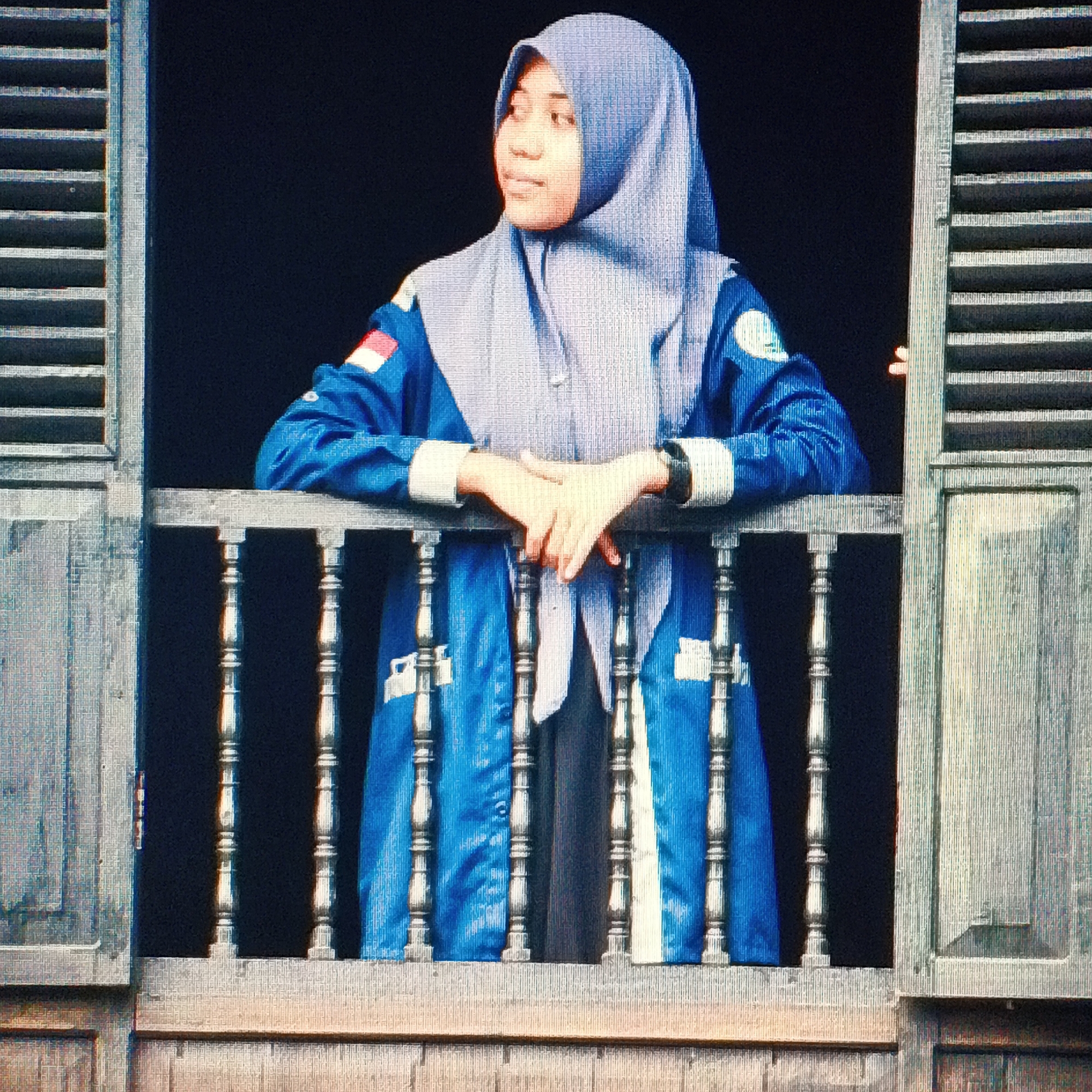 A medium shot shows a young Indonesian woman wearing a light blue hijab and a blue jacket with white accents and emblems on the sleeves. She is standing at a railing, leaning on it with her crossed arms. The railing consists of turned wooden balusters and a top rail. She is framed by an open window with weathered wooden shutters on the left and right. The background is a dark, solid color, suggesting an interior space or a very dark exterior. The image appears to have a textured or pixelated quality. The woman's expression is serene as she looks to her left. The scene is likely an indoor setting looking out, or a shaded outdoor area. Without additional context, a specific location within Ende, Indonesia cannot be determined.