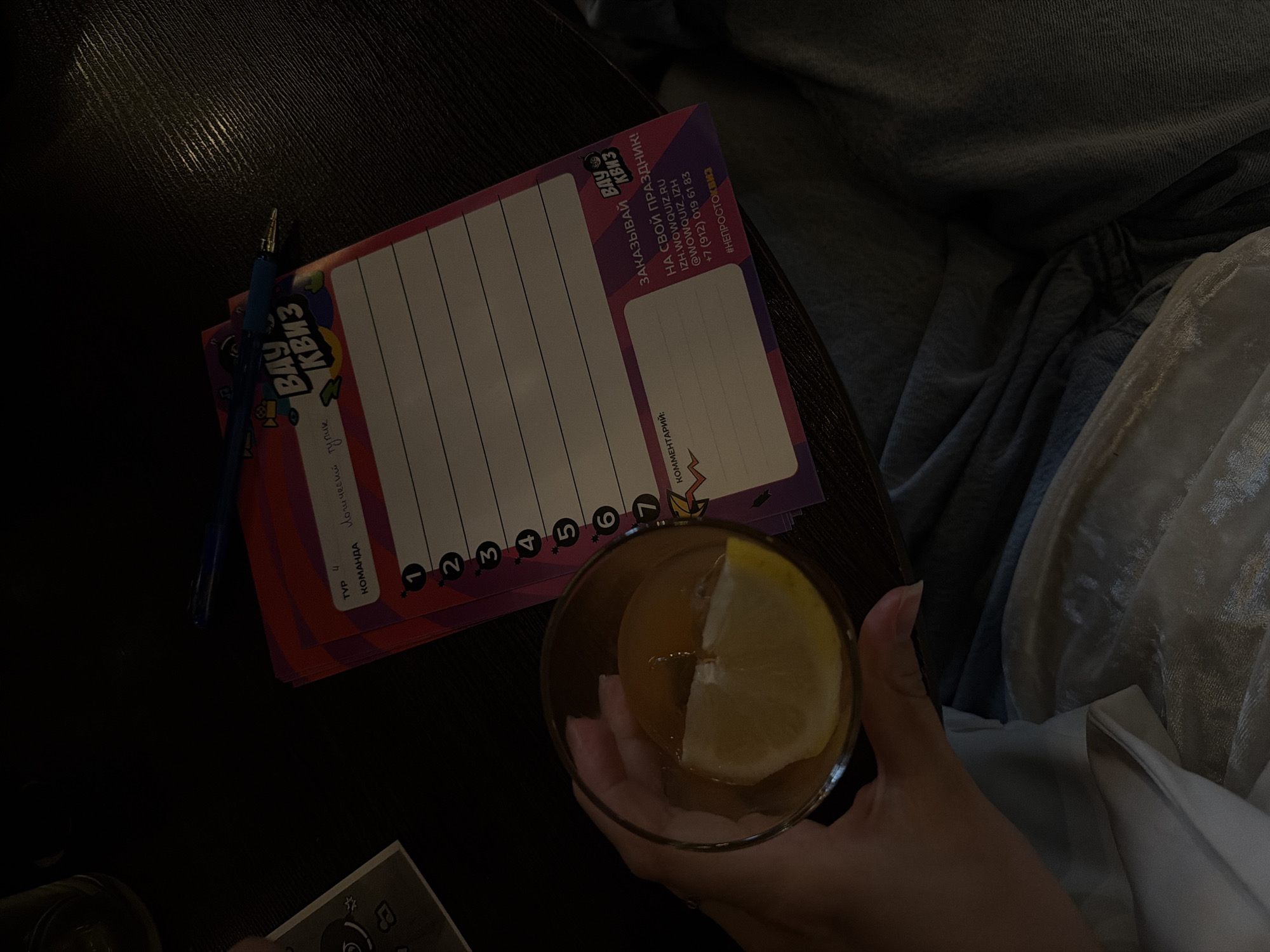 The image shows a cocktail with a lemon slice held in a person's hand next to a "Vau Quiz" trivia card from Izhevsk, Russia. The card is numbered 1 through 7 for answers and has a space for comments. A pen is resting near the top of the card. The background includes a dark surface, possibly a table, and the edge of clothing or a blanket.