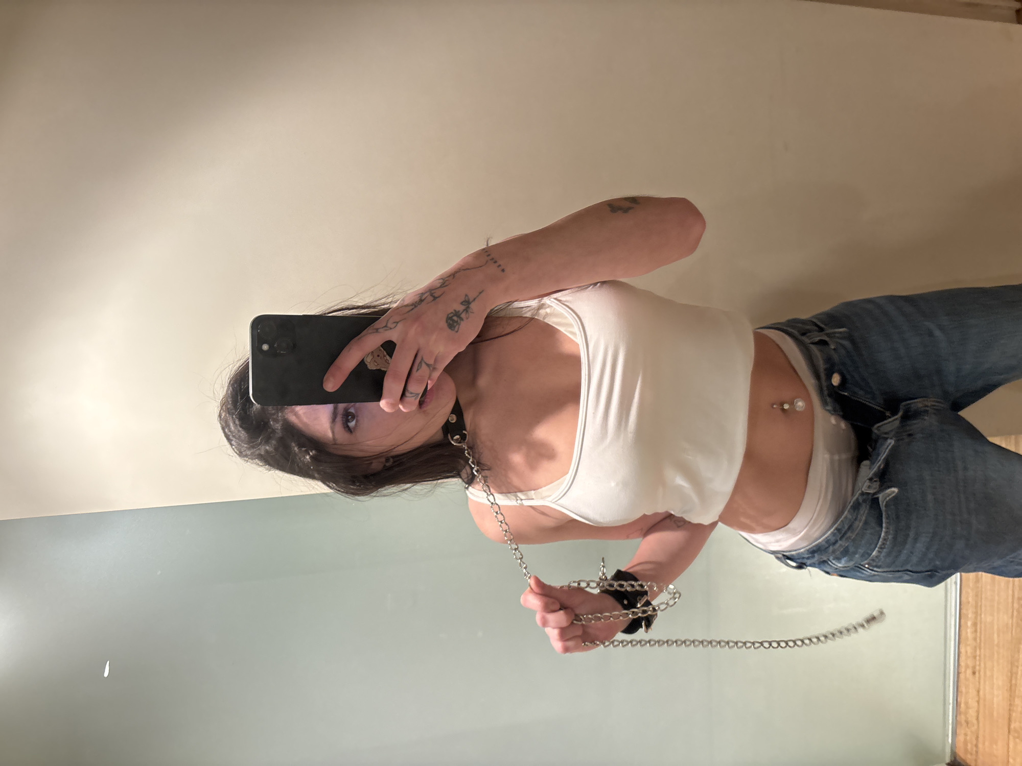 An indoor mirror selfie taken in Buenos Aires, Argentina, features a person, likely female, standing upright. They are dressed in a white sleeveless crop top, blue denim jeans, and a white undershirt or underwear peaking above the jeans' waistband. A belly button piercing with two clear gems is visible on their midriff. The person holds a dark-colored smartphone, likely an iPhone, up to capture their reflection, partially obscuring their face; one eye is visible looking towards the camera.
Visible on their right forearm and hand are several distinct tattoos, including floral or vine-like designs and some indistinct text. They wear a black choker or collar around their neck, and a black cuff on their right wrist. Silver-colored chains connect the collar and wrist cuff, extending downwards and held in the person's hands. The background is simple, with a light beige or cream-colored wall in the upper portion and a reflective, light blue-grey surface below, possibly a mirror or a glossy wall. The lighting suggests an interior setting.