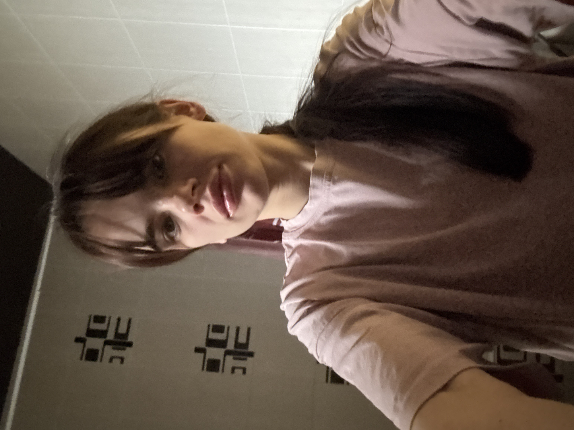 A woman with brown hair and bangs is facing the camera. She is wearing a light pink t-shirt. The background features a tiled wall with a repeating geometric pattern and a white tiled ceiling. Based on the tiling, this image may be taken inside a bathroom, but it is not possible to confirm if the image was taken in the city of Rostov-on-Don. No specific actions or interactions are apparent in the image.