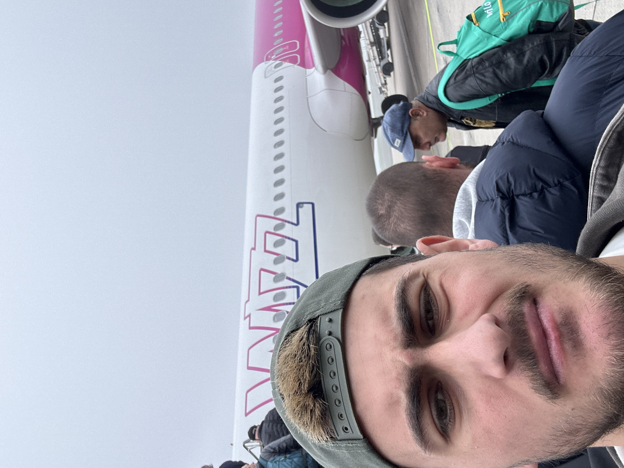 The image shows a man in the foreground, wearing a green baseball cap and a light jacket. Behind him is an aircraft, identifiable by the pink and purple "WIZZ" logo on its tail. Other individuals are seen in the background, suggesting a group of people boarding or deboarding. The environment is overcast, with a gray sky visible. Given the presence of the "WIZZ" logo, it's plausible the location is near or at Dortmund Airport in Dortmund, Germany, as Wizz Air operates flights to and from this airport. However, the exact location within Dortmund cannot be definitively confirmed from the image alone.