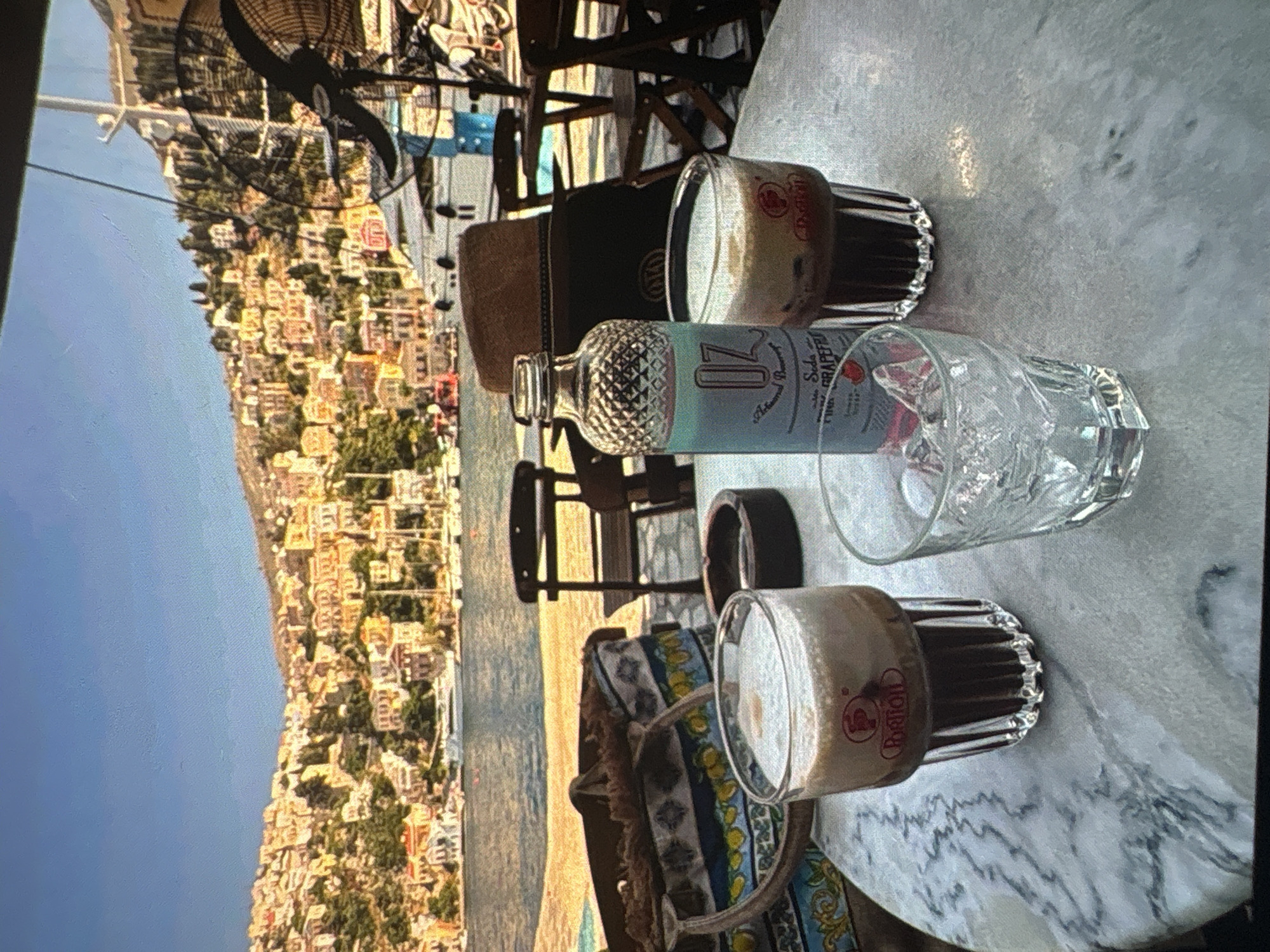 The image, originally rotated, depicts an outdoor cafe or restaurant setting overlooking a scenic coastal town and the sea. The foreground features a round, light-colored marble table laden with drinks. Two glasses, characterized by a ribbed design, contain a dark liquid with a foamy top, resembling iced coffee. One of these glasses clearly shows red Greek text, "Ραδάλ", which also appears as part of a circular logo. An empty clear glass filled with ice cubes sits next to a distinct glass bottle with a textured, patterned top half and a light blue label. Visible text on the label includes "17", "SODA", "GRAPEFRUIT", and "PINK", along with partially visible words like "Artisanal" and "Beverage". Beneath the table or on an adjacent chair, parts of a dark brown bag and a patterned cushion (blue, yellow, white) are visible. A black fan is also partially visible in the upper part of the foreground.
Beyond the dining area, a clear blue body of water, presumably the sea, stretches into the midground. Several small boats can be faintly seen on the water. The background showcases a densely populated hillside, with numerous light-colored buildings and houses cascading down towards the coastline. Green foliage is interspersed among the structures, suggesting a lush landscape. The sky above is clear and blue, indicating bright, sunny daytime weather. The scene conveys a relaxed, leisurely atmosphere, likely set in a Mediterranean location given the architecture and the Greek text on the glasses.