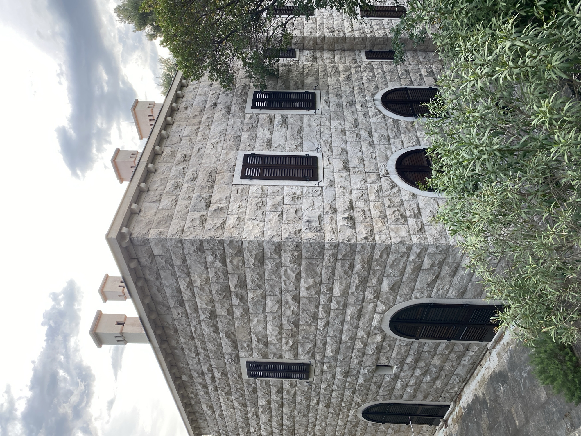 An upward-angle shot captures a stone building in Sveti Stefan, Montenegro. The building is made of gray brick and features a row of small, tower-like structures along the top edge. Windows with dark shutters, some rectangular and some arched, are visible. A tree with green leaves is present on the right side of the frame. A cloudy sky is partially visible on the left.