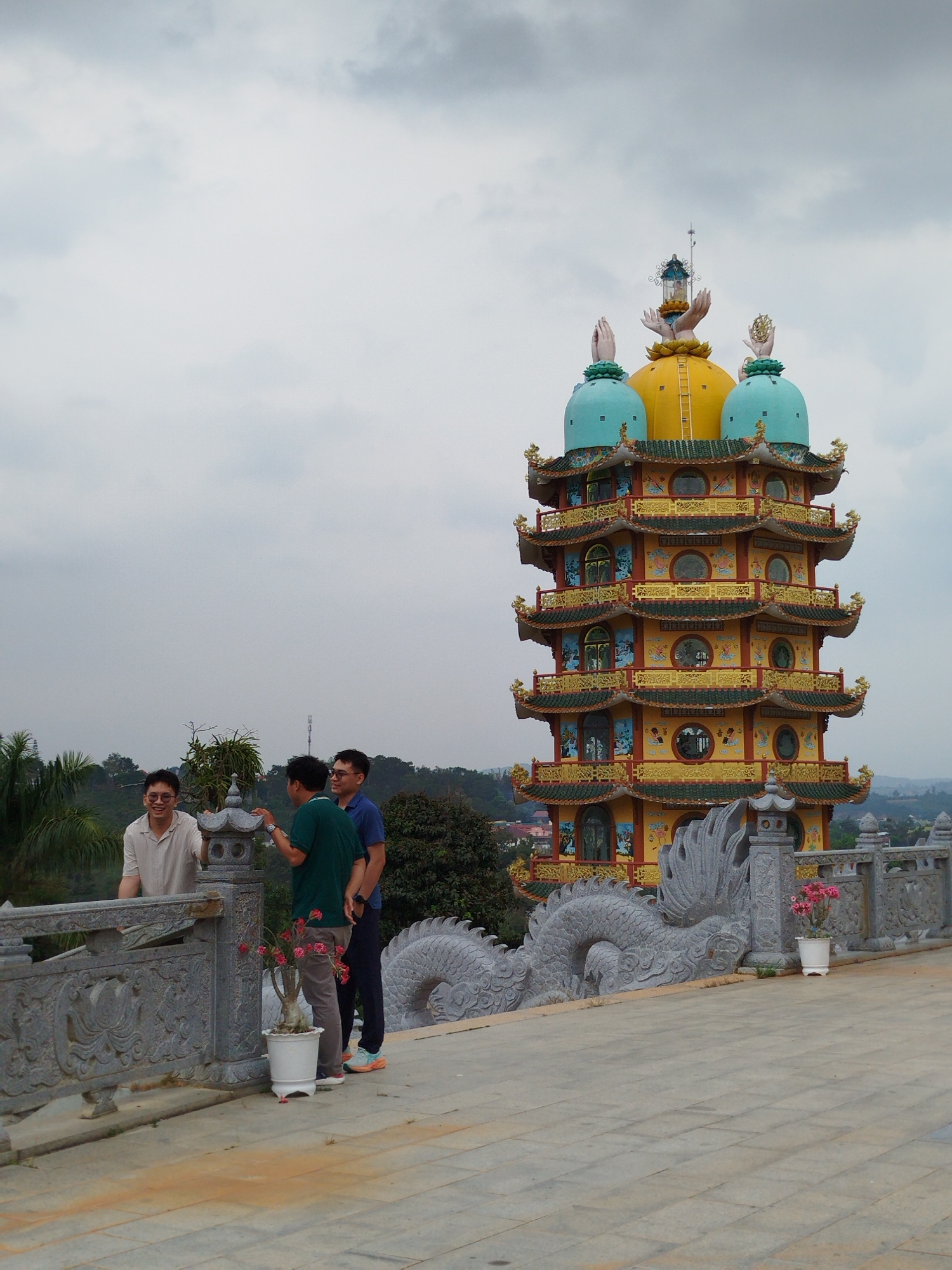 Three men stand on a stone platform near a pagoda-style structure. The pagoda is tall, multi-tiered, and brightly colored with yellow, gold, and blue accents. A decorative stone dragon runs along the edge of the platform. The men are casually dressed and appear to be conversing. The background shows a cloudy sky and a somewhat distant view of trees and possibly other buildings. The location is Thiện Chí, Vietnam.