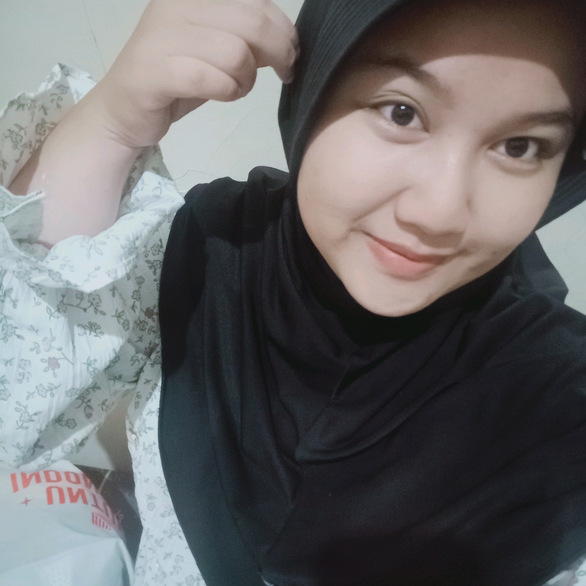 The image is a close-up selfie of a young woman wearing a black hijab and a white floral-patterned blouse. She is indoors, with a plain, light-colored wall in the background. In the lower left corner of the image, a white plastic bag with red text that appears to be "INDO" and "PI" is visible. The woman is looking at the camera and smiling. Her left arm is bent upwards, and she is touching the edge of her hijab with her fingers. The lighting in the image is soft, suggesting it was taken in natural light or with indoor lighting. The overall scene appears to be casual and personal.