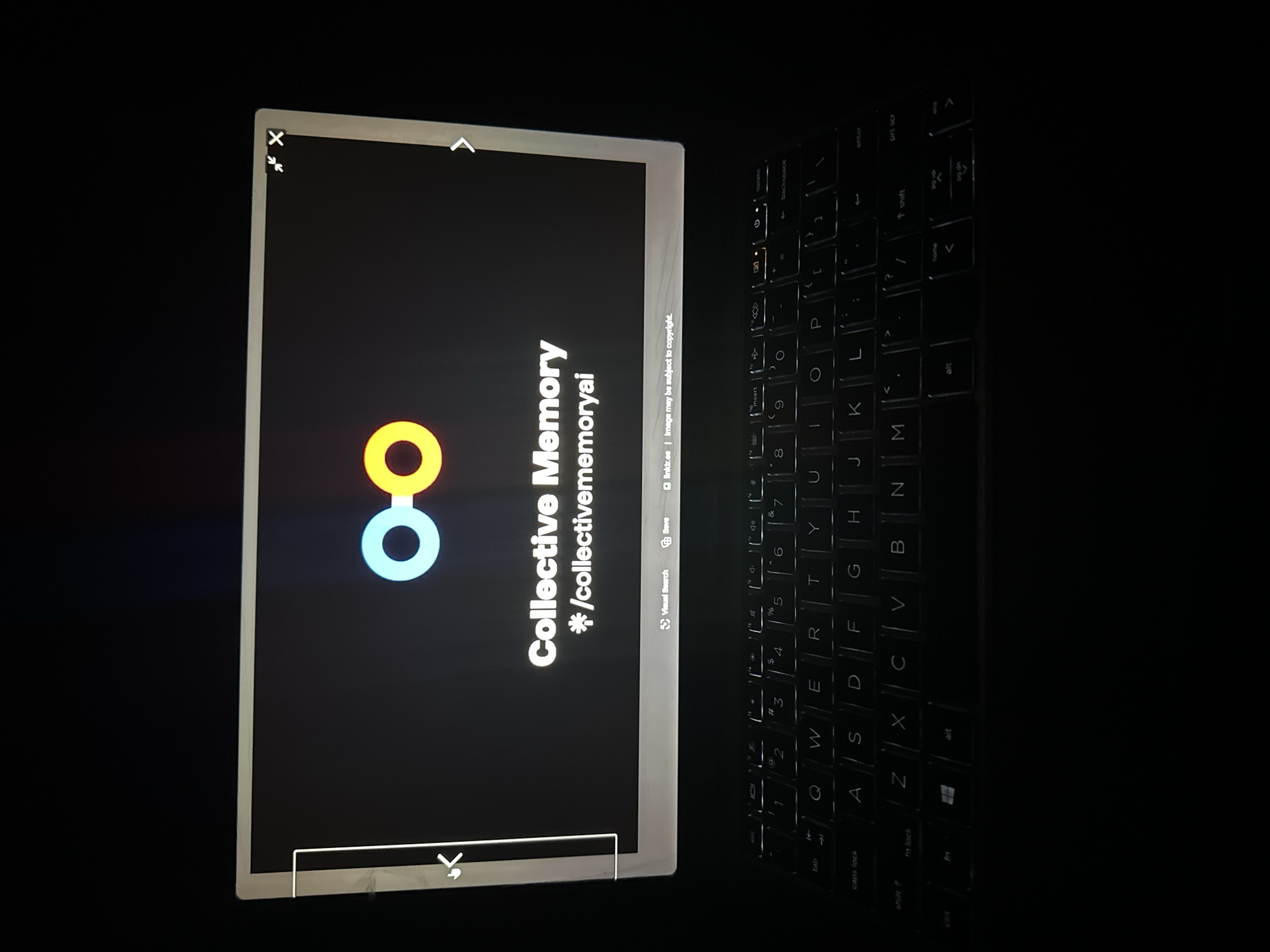The image displays a laptop computer in what appears to be a dimly lit or dark environment, suggesting nighttime or an indoor setting with minimal lighting. The image is rotated 90 degrees clockwise, showing the laptop vertically.
The primary subject is the laptop's screen, which presents a black background with a prominent logo and text. The logo consists of two interconnected or overlapping circular shapes: one orange/yellow and one light blue. To the right of the logo, the text "Collective Memory" is displayed in large white letters, followed by "*/collectivememoryai" on the line below. At the bottom of the screen's content area, there's a small section with "linktr.ee Image may be subject to copyright." Below the screen's viewing area, part of the application's interface shows buttons labeled "Visual Search" and "Save." The top right corner of the screen has common UI elements: an "X" (close), an upward-pointing caret (maximize/restore), and a downward-pointing caret (minimize).
To the right of the screen, a portion of the laptop's backlit QWERTY keyboard is visible. Individual keys are illuminated, showing letters, numbers, and symbols such as "Q", "W", "E", "R", "T", "Y", "U", "I", "O", "P", "A", "S", "D", "F", "G", "H", "J", "K", "L", "Z", "X", "C", "V", "B", "N", "M", along with function keys, "tab", "caps lock", "shift", "alt", "enter", "backspace", "delete", "prt scr", "home", and "insert". The laptop has a light-colored, possibly silver or white, bezel around the screen.