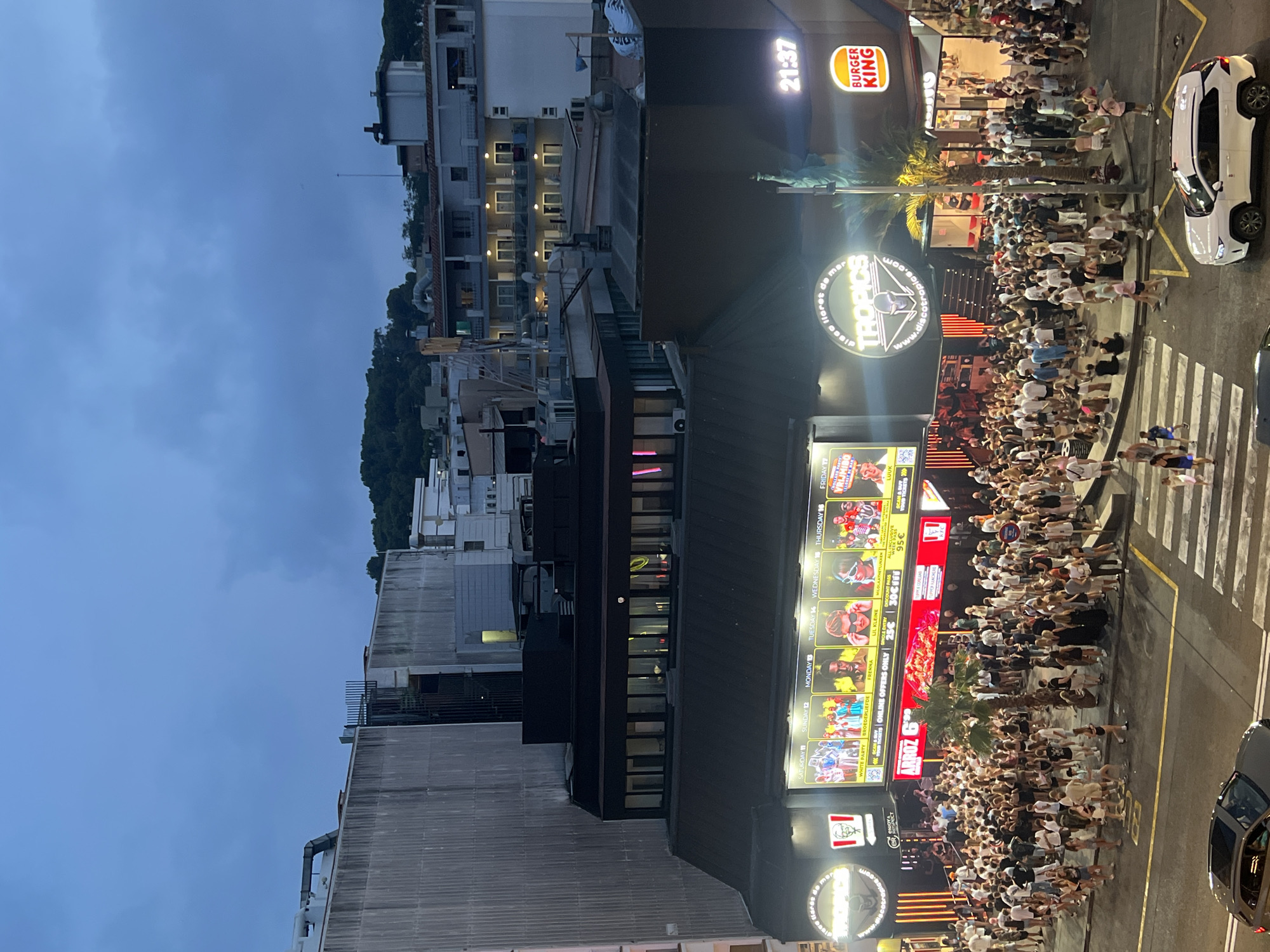 The image shows a street scene in Lloret De Mar, Spain, at dusk or early evening. A large crowd of young people, dressed in casual summer clothing, are gathered outside a nightclub called "Tropics" and adjacent establishments including a KFC and a Burger King. The nightclub features a large digital billboard displaying advertisements for various events and performers. The street is busy with pedestrians, and several cars are visible on the road, including a white SUV. Palm trees are present, indicating a coastal or warm climate. The overall atmosphere appears lively and social, typical of a popular entertainment district.