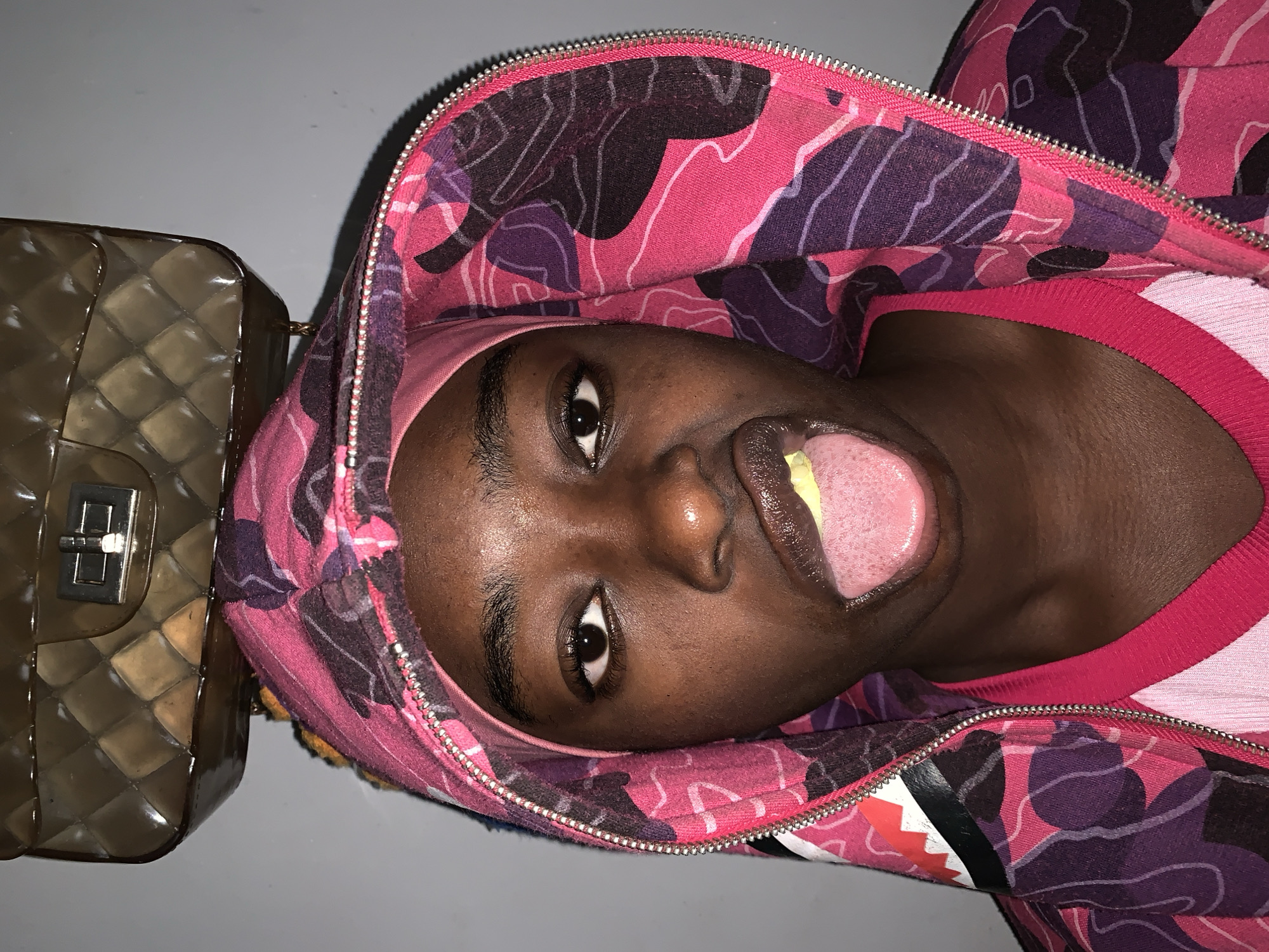 A person with dark skin is wearing a pink and purple camouflage zip-up hoodie and sticking their tongue out. The person is wearing a pink headband and a pink shirt underneath the hoodie. A clear, quilted purse with a silver clasp is positioned to the left of the person's head. The background appears to be a plain, light-colored surface. The image is oriented at an angle. The image does not show Rafin Par.