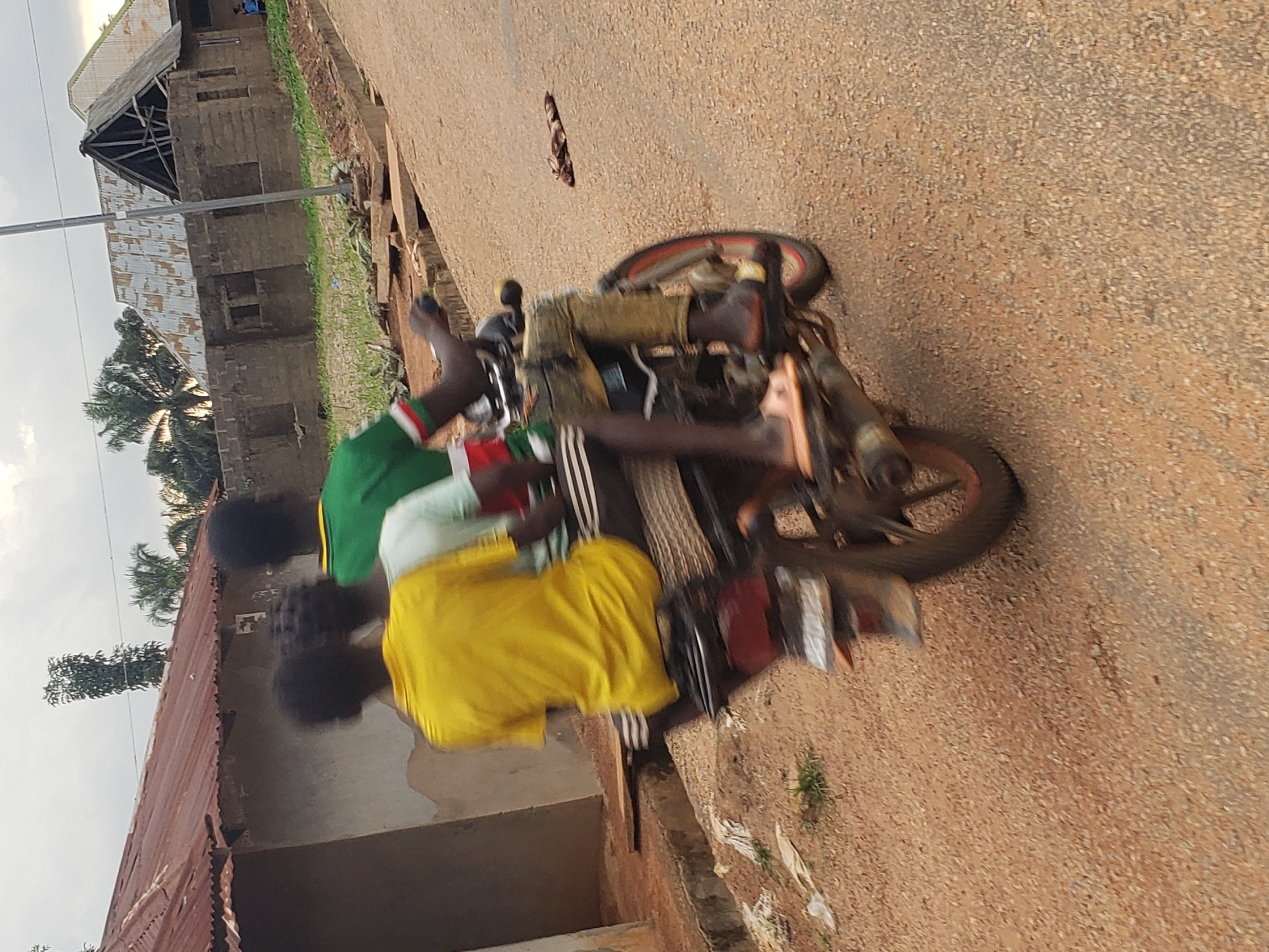 In Kachia, Nigeria, a motorcycle with three young individuals is captured in motion on a dusty, unpaved road. The person in the driver's seat is wearing a green and white striped shirt, black pants, and is holding the handlebars. Directly behind the driver sits another person wearing a bright yellow t-shirt and black pants with white stripes. The third passenger, positioned behind the person in the yellow shirt, is partially obscured but appears to be wearing a light-colored shirt. The motorcycle is a dark color with red detailing on the wheels. The surrounding environment includes rudimentary buildings with corrugated metal roofs and block walls, indicative of a developing urban or semi-rural setting. Vegetation is sparse, with some small plants growing along the roadside. The overall impression is one of everyday life and transportation in this Nigerian city.