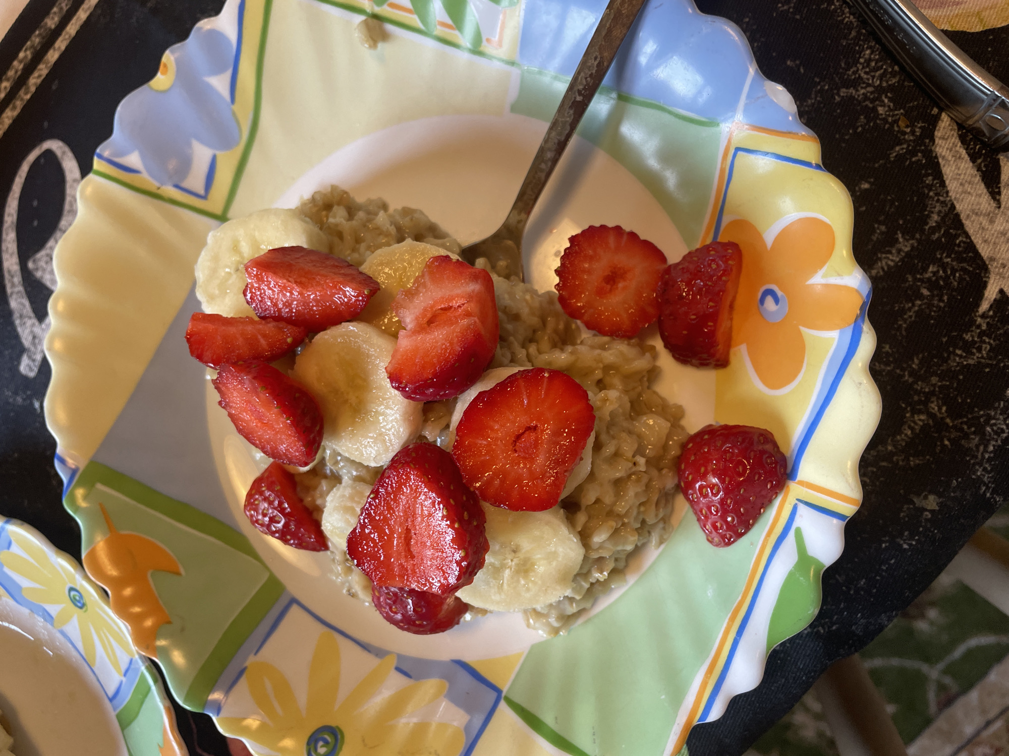An overhead shot shows a bowl of oatmeal topped with sliced strawberries and bananas. The bowl sits on a decorative plate with a floral pattern. A fork rests on the plate to the right of the bowl. The scene appears to be indoors, likely in a home setting, as indicated by the presence of a table and tableware. There is no identifiable context to place this image in the city of Турово, Russia.
