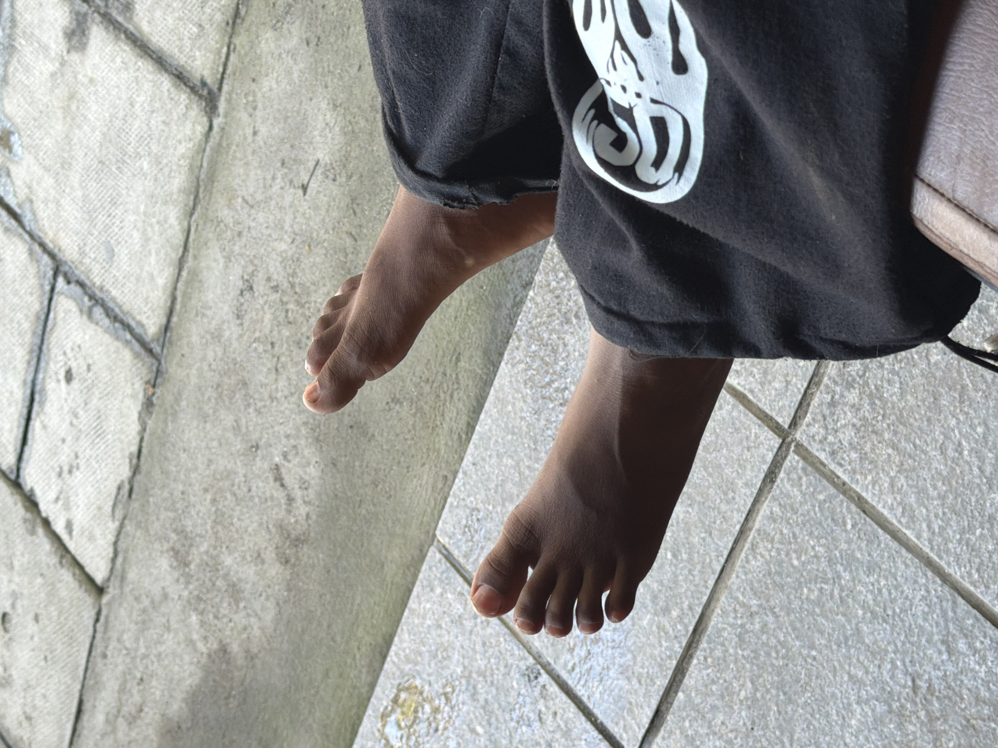 The image shows a close-up view of a person's bare feet resting on a tiled floor. The person is wearing black pants with a white graphic design on them. The tiled floor appears to be outdoors, possibly in a public space or residential area. The lighting suggests it is daytime. The location is identified as Mengwi, Indonesia.
