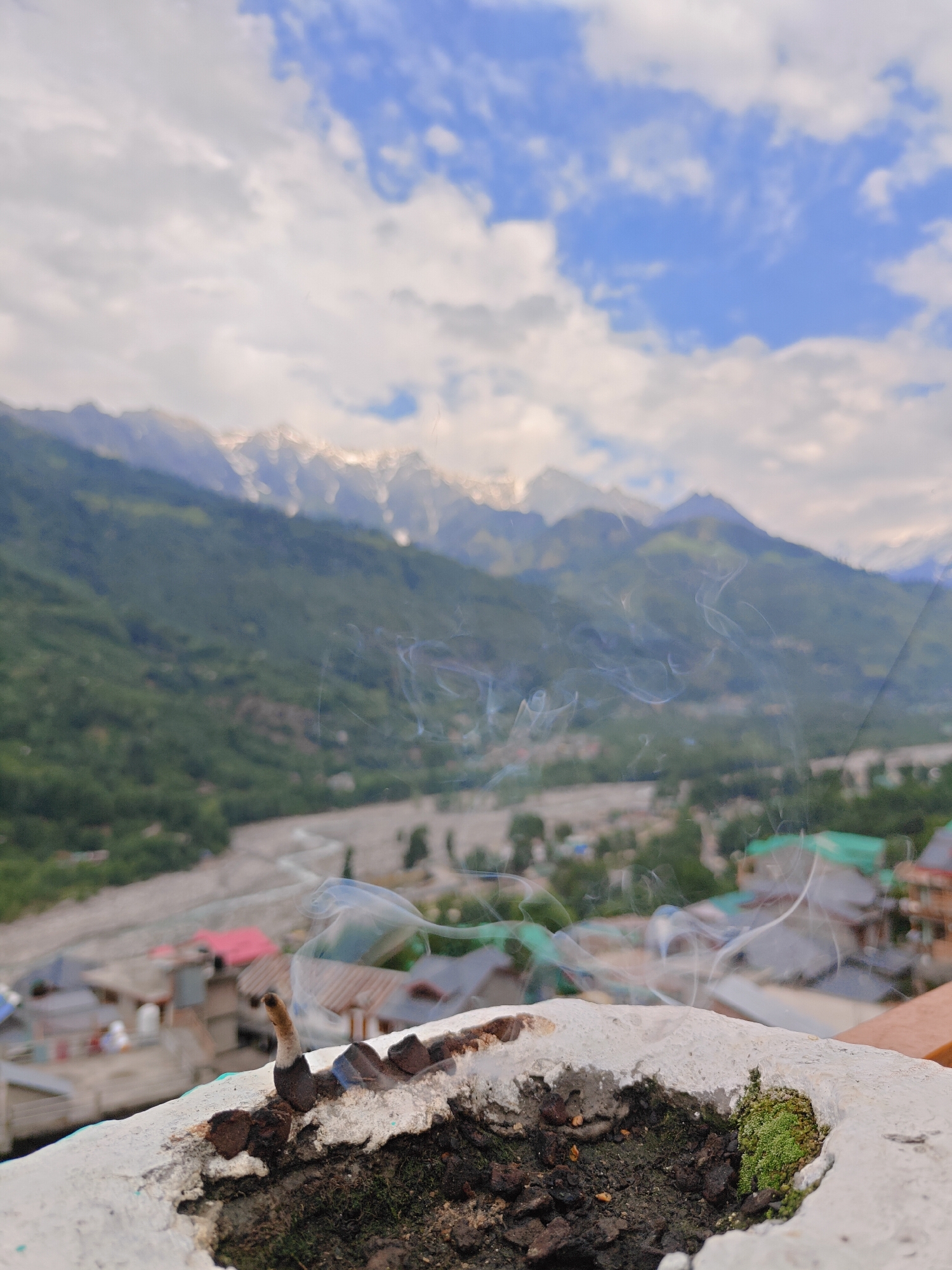 The image captures a scenic view of the Himalayas from Vashisht, India. In the foreground, a white ledge or wall is visible, with moss and burnt material on it. A single incense stick is burning, emitting wisps of smoke that drift upwards and across the frame. Beyond the ledge, a village with traditional Indian architecture, featuring red and green rooftops, is nestled in a valley. A river flows through the valley, and the slopes of the mountains are covered in lush green trees. In the background, the majestic snow-capped peaks of the Himalayas pierce the cloudy blue sky, with sunlight illuminating their summits. The overall scene evokes a sense of peace and natural beauty.