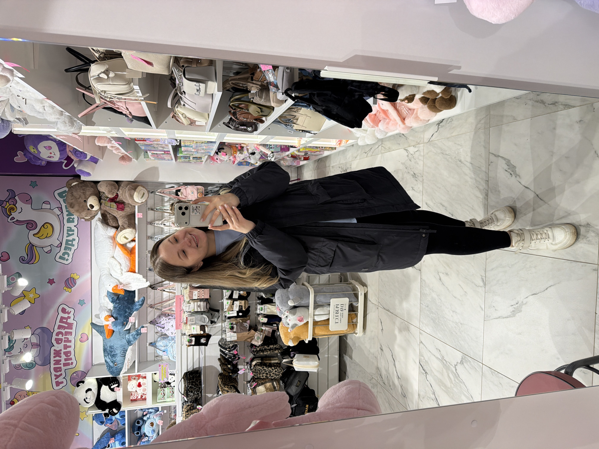 The media file captures a mirror selfie taken by a young woman inside a brightly lit retail store. The image is presented rotated 90 degrees clockwise, but when viewed upright, it shows the woman smiling at her smartphone as she holds it up to capture her reflection. She has long, light brown hair and is wearing a black long coat over a light blue top, black leggings, and white sneakers.
The setting is an indoor retail space, likely a gift shop or accessory store, characterized by modern white shelving units displaying a wide array of merchandise. These include numerous handbags and backpacks in various colors (white, pink, beige, black), as well as a large collection of plush toys. Visible plush animals include a brown teddy bear, a purple character, a white goose, blue sharks, a panda, and a blue Stitch figure. Smaller stuffed animals are placed in a clear plastic bin, and folded blankets or throws are visible in a basket. Other shelves hold various socks, headbands, and small accessories.
The floor is covered with elegant white and grey marble-patterned tiles. A colorful wall decal featuring unicorns, stars, and candies is prominent on the left side of the store. Bright overhead spotlights illuminate the space. Part of a pink chair with a black base is visible in the foreground.
Visible Cyrillic text on the wall decal clearly reads "ЗДЕСЬ ЖИВУТ ЧУДЕСА," which translates to "HERE LIVE MIRACLES." Another sign on a product bin containing blankets reads "ДЛЯ ДОМА," meaning "FOR HOME." The woman's joyful expression and the store's cheerful decor suggest a pleasant shopping experience in a modern retail environment, consistent with a location like Izhevsk, Russia.