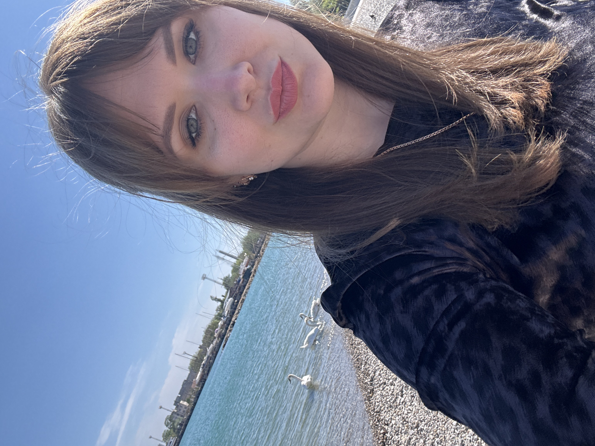 A selfie image shows a young woman with light brown hair and green eyes. She is wearing a dark-colored shirt. The background reveals a body of water, likely a lake or sea, with several swans swimming in it. The shoreline consists of pebbles. In the distance, buildings and masts are visible, suggesting the location might be a waterfront area with a harbor. Although I cannot specify the exact location within Yevpatoriya, Ukraine, based on the visual data, the scene resembles a coastal area with architectural structures and water-related activity in the background.