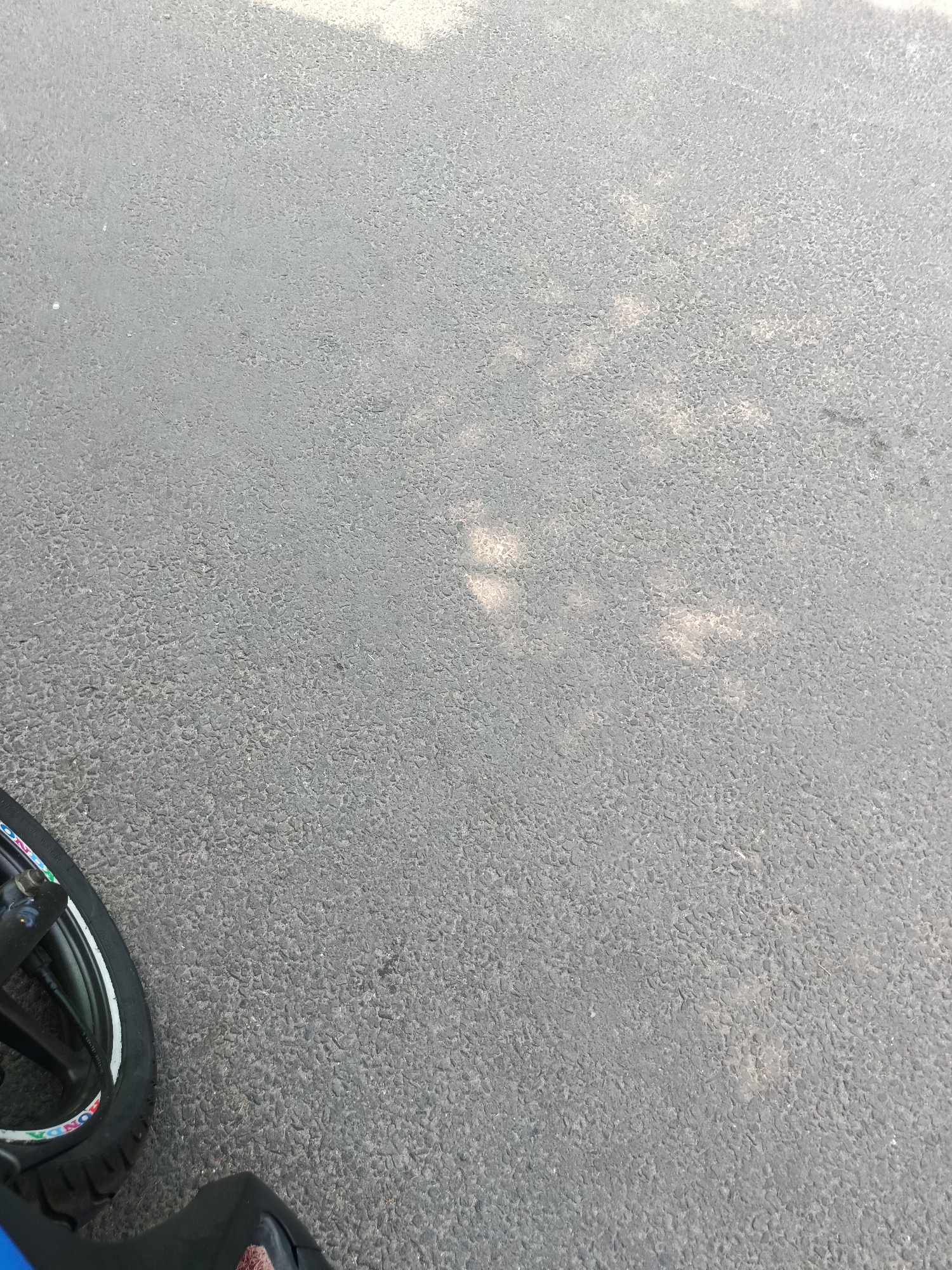 The image displays a close-up view of asphalt pavement, likely a road surface. The texture of the asphalt is granular and appears to be in good condition. There are dappled shadows cast across the surface, suggesting it is outdoors and illuminated by sunlight, possibly filtered through trees or other overhead structures. In the bottom left corner, a portion of a black tire with white and colored decals is visible, indicating the presence of a vehicle, likely a motorcycle or bicycle, partially captured in the frame. A dark blue object, possibly part of the vehicle's body, is also present at the very bottom of the image. The overall scene is static and focuses on the ground surface. No specific landmarks or buildings are discernible, making it difficult to pinpoint the exact location. However, if this image is indeed from East Jakarta, Indonesia, it represents a common urban streetscape.
