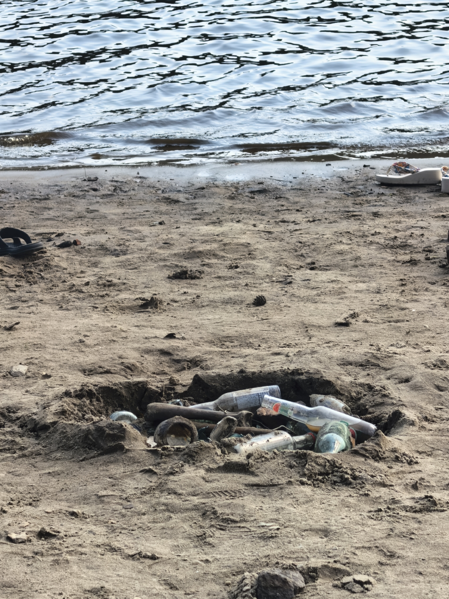 The image depicts a sandy shore of a body of water, likely a river or lake, as indicated by the rippling surface in the background. The foreground is dominated by sand with some scattered debris. Notably, there is a pit in the sand filled with several glass bottles, some of which are partially buried. Additionally, a pair of black flip-flops can be seen on the sand to the left, and a pair of white flip-flops are visible on the right. The overall scene suggests a location in the city of Sysert, Russia, perhaps a recreational area that has been littered. There are no people visible in the image.