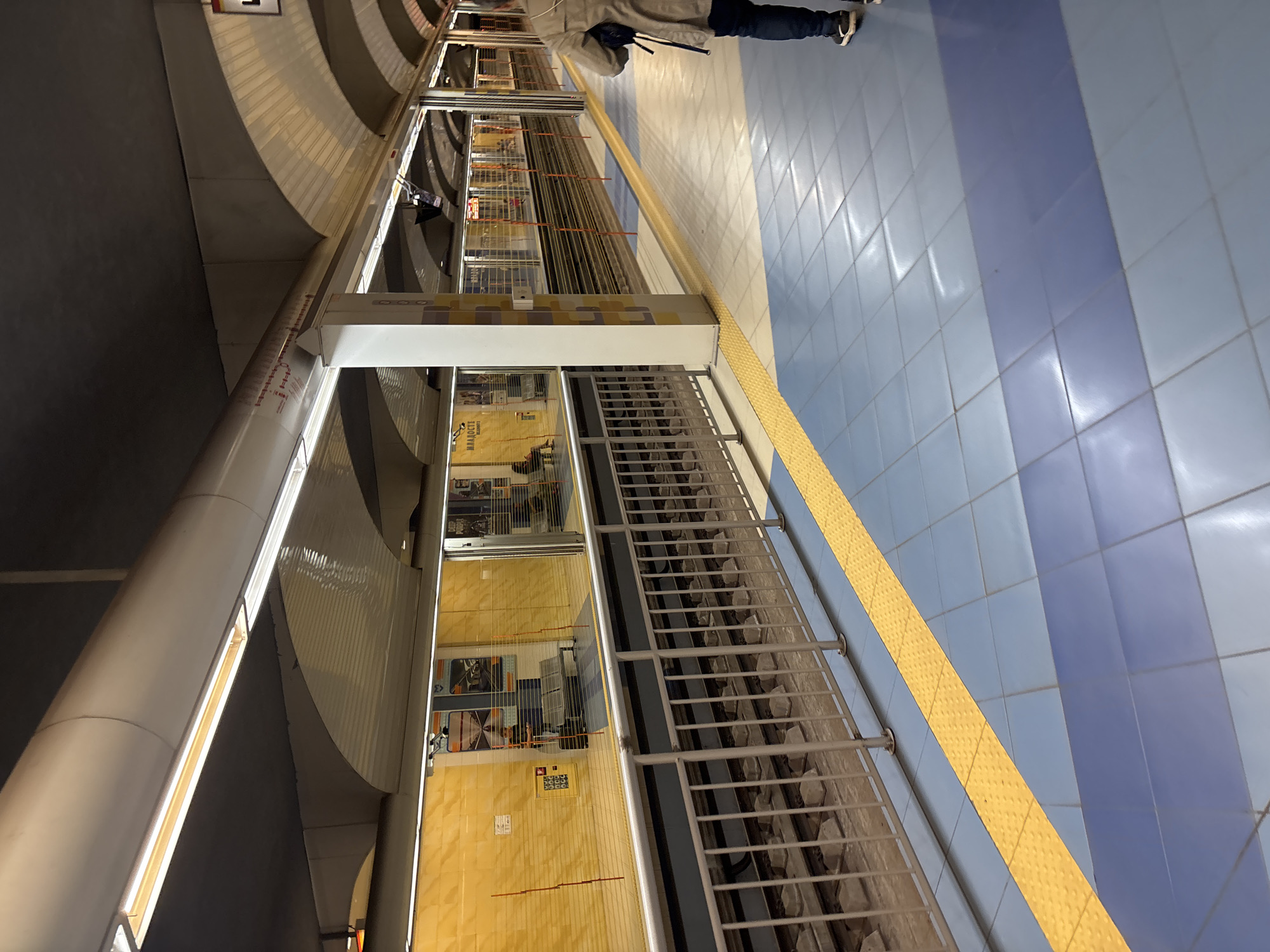 This image shows a station of the Sofia Metro in Sofia, Bulgaria. The platform is tiled in a light blue and dark blue pattern, and a yellow tactile paving strip runs along the edge. A train is visible behind a glass safety barrier. A person wearing a coat and blue jeans is standing on the platform.