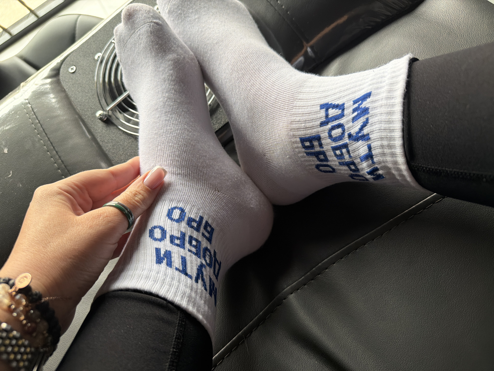 A close-up shot shows a person's legs and feet resting on what appears to be a black leather or vinyl surface. The individual is wearing white socks with blue Cyrillic text on the ankles. The text seems to read "Мути Добро Бро", which translates to "Make Good Bro". The person is also wearing black leggings.
The person's hand is visible on the left side of the image, with manicured nails and a ring on one finger. A beaded bracelet is also on the wrist.
Behind the feet and legs, there is a piece of equipment with a fan grill, suggesting an environment like a nail salon or spa. The image doesn't provide enough specific detail to determine the city or region definitively. The location could be Unknown, Unknown, but there is no way to confirm this with only the given image.