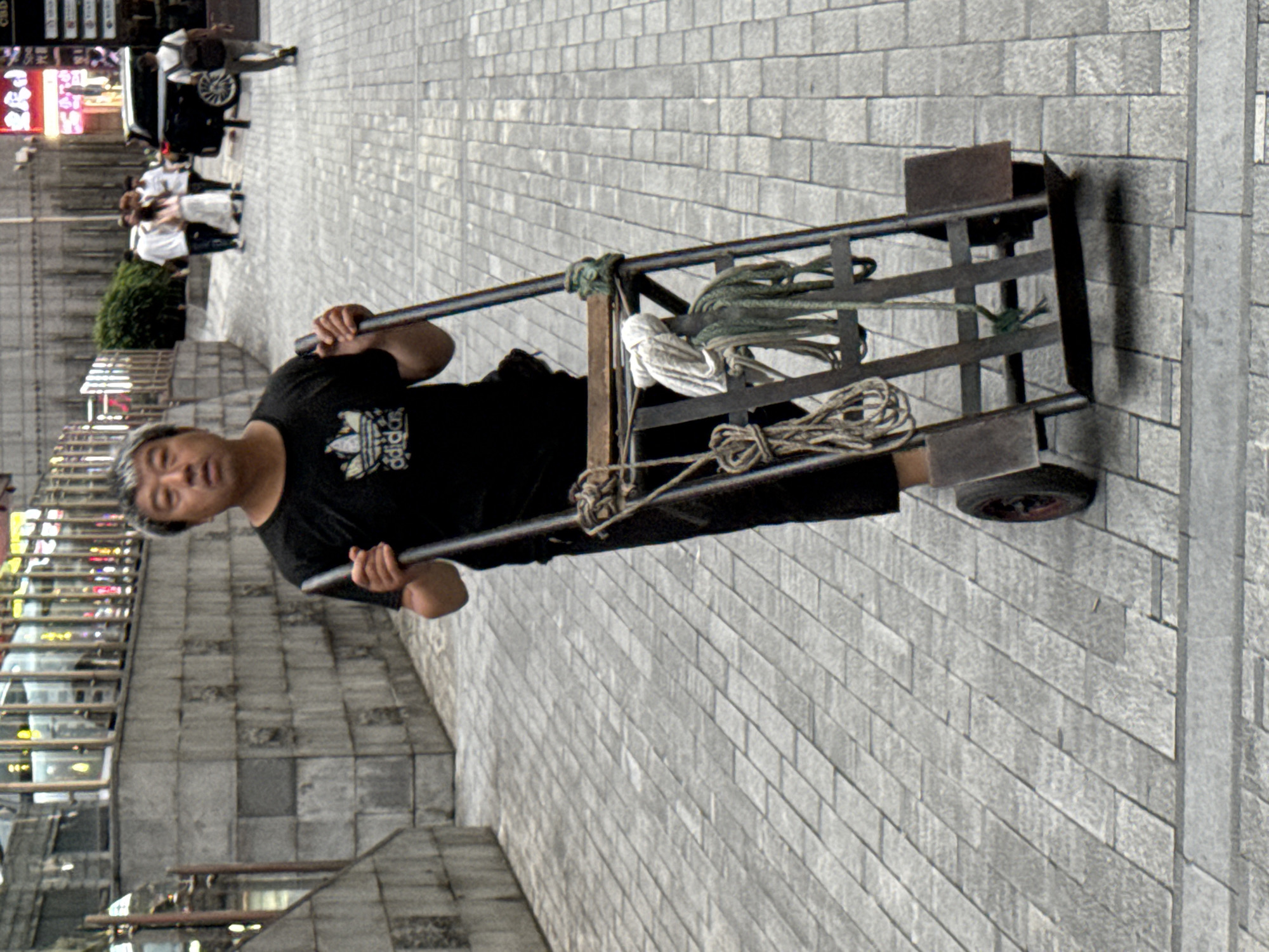 A middle-aged East Asian man with graying hair is pushing a metal handcart down a sidewalk paved with gray rectangular stones. He is wearing a black t-shirt with the Adidas logo and black pants. The handcart has two wheels and appears to be loaded with bundles of rope and a white cloth. In the background, several other people are walking, and a black car is parked on the street. The scene is likely in a commercial area of Yuzhong District, China, given the presence of buildings with illuminated signs and the architectural style of the sidewalk. The lighting suggests it is daytime, possibly overcast.