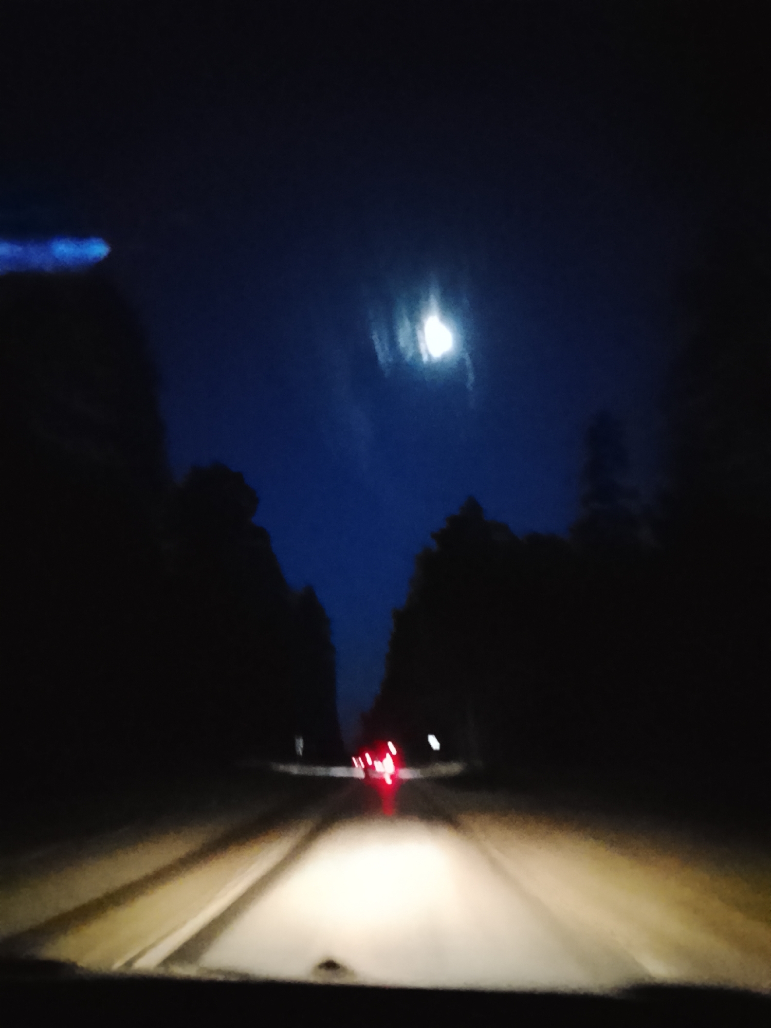 A nighttime view from the interior of a vehicle traveling on a road. The road is illuminated by the vehicle's headlights. Dense, dark trees flank both sides of the road, suggesting a rural or wooded area. The sky is visible above the trees, with a bright moon and a few faint clouds. Further down the road, the red taillights of another vehicle can be seen.
The location is Высокиничи, Russia.