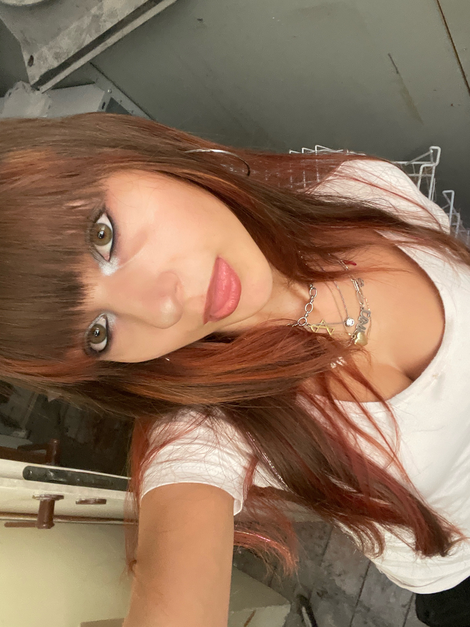 The image shows a young woman with long, dark brown hair with reddish highlights, wearing a white t-shirt and layered silver necklaces. She is looking at the camera with heavy eye makeup. The background appears to be an indoor industrial or unfinished space, with concrete walls and some metal structures visible. The image is a selfie, taken from a slightly tilted angle. The image is oriented vertically. There is no indication of the city of Raanana, Israel within the image or its context.