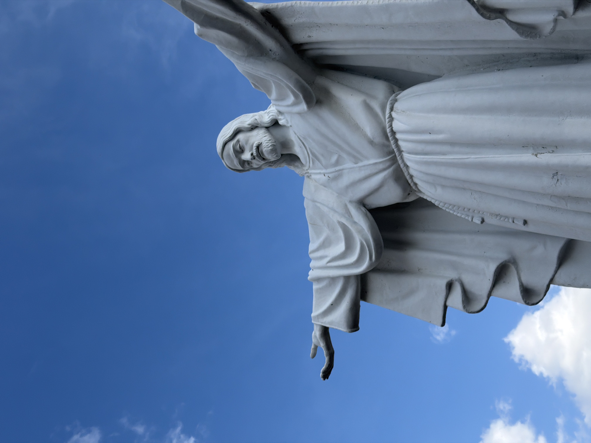 The image shows a statue of Jesus Christ against a blue sky with a few scattered white clouds. The statue is white, depicting Jesus in flowing robes with outstretched arms. A rope belt is visible around his waist. The statue is located in Krasnoyarsk, Russia.