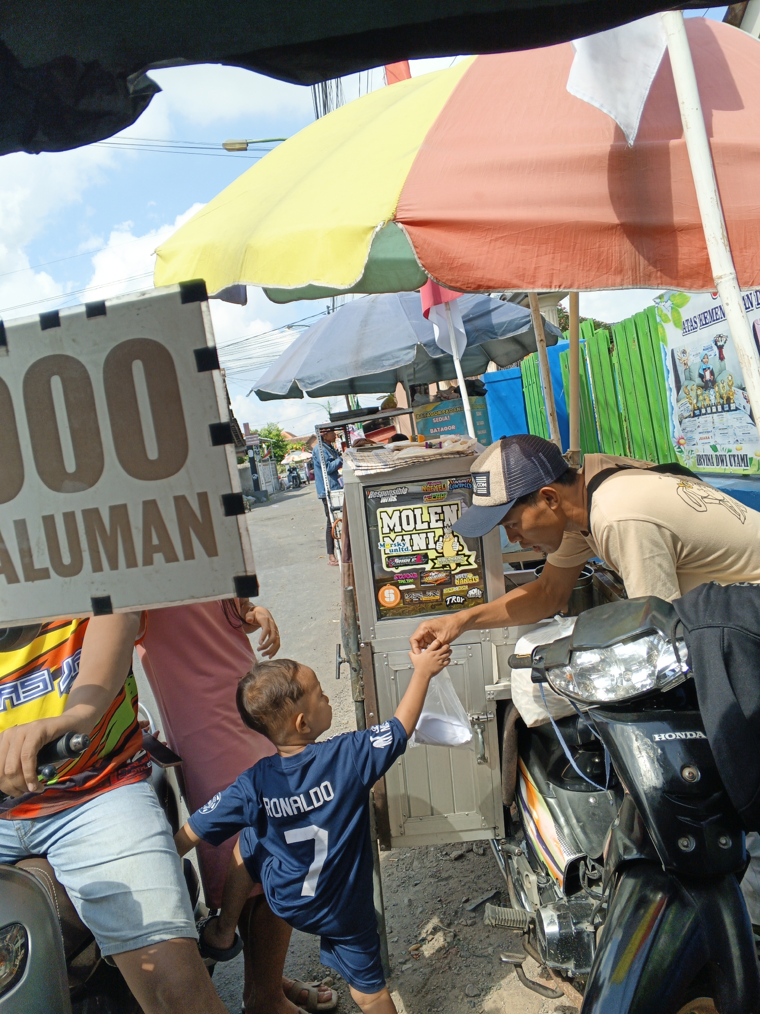 In Padang, Indonesia, on a sunny day, a young boy wearing a blue "RONALDO 7" soccer jersey is handing a plastic bag to a man. The man, wearing a beige t-shirt and a baseball cap, is leaning over to receive the bag from the boy. They are standing next to a mobile food cart, which has colorful signage and appears to be selling "Molen Mini." A black motorcycle is parked to the right of the cart, with its headlight illuminated. In the background, a street is visible with other vendors and a green fence. The scene is partially covered by large, colorful umbrellas.