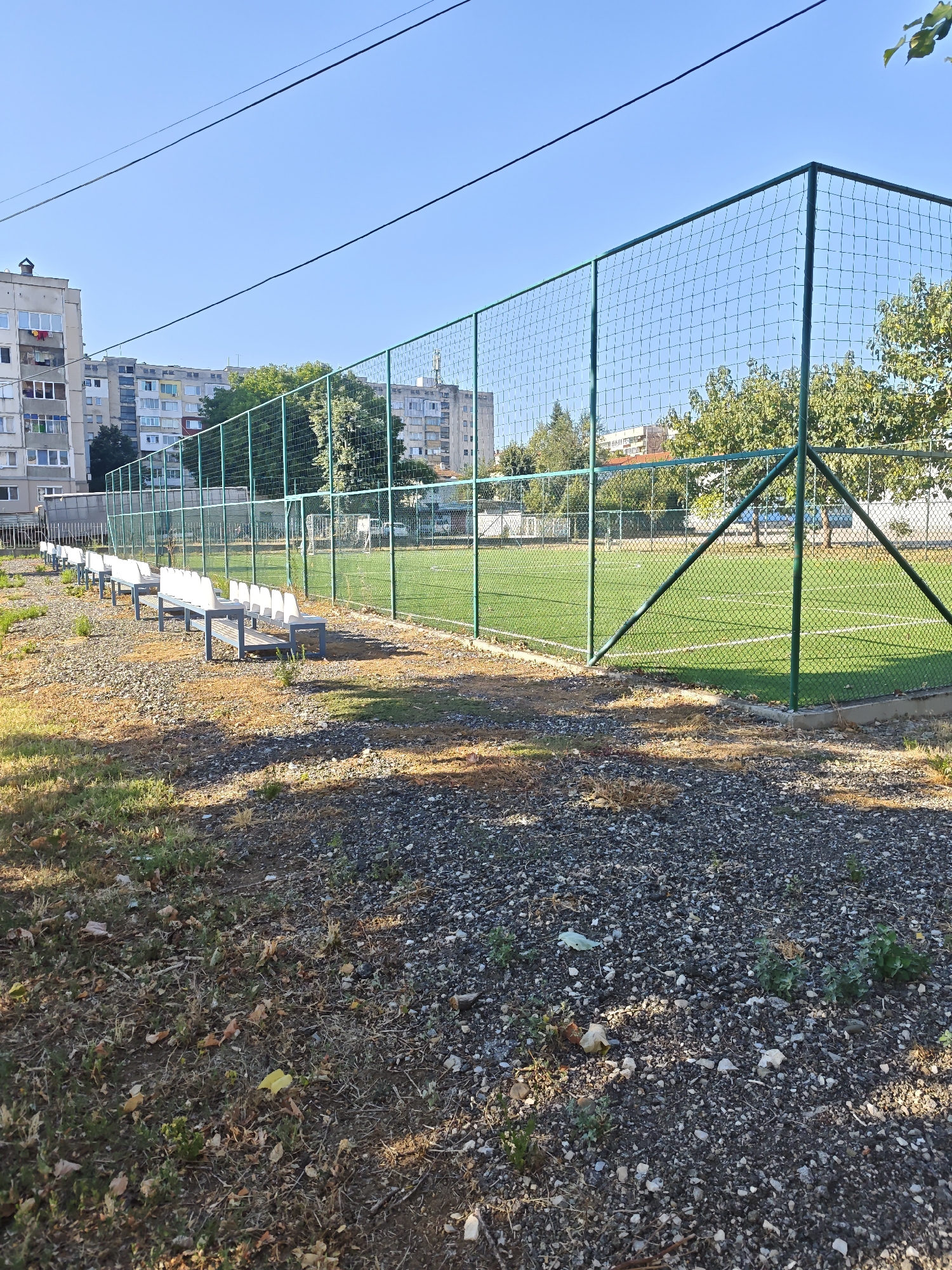 The image shows an outdoor recreational area with a fenced-in artificial turf sports field, possibly a small soccer pitch. The field is marked with white lines. In the foreground, a row of white plastic benches with grey metal legs is situated on a gravel surface covered with dry leaves and sparse vegetation. A green metal fence encloses the sports field. Beyond the fence, trees are visible, and further in the background, several multi-story residential buildings can be seen under a clear blue sky. The overall scene suggests a public park or a school sports facility. The location is identified as being in Lovech, Bulgaria. There are no people visible in the image.