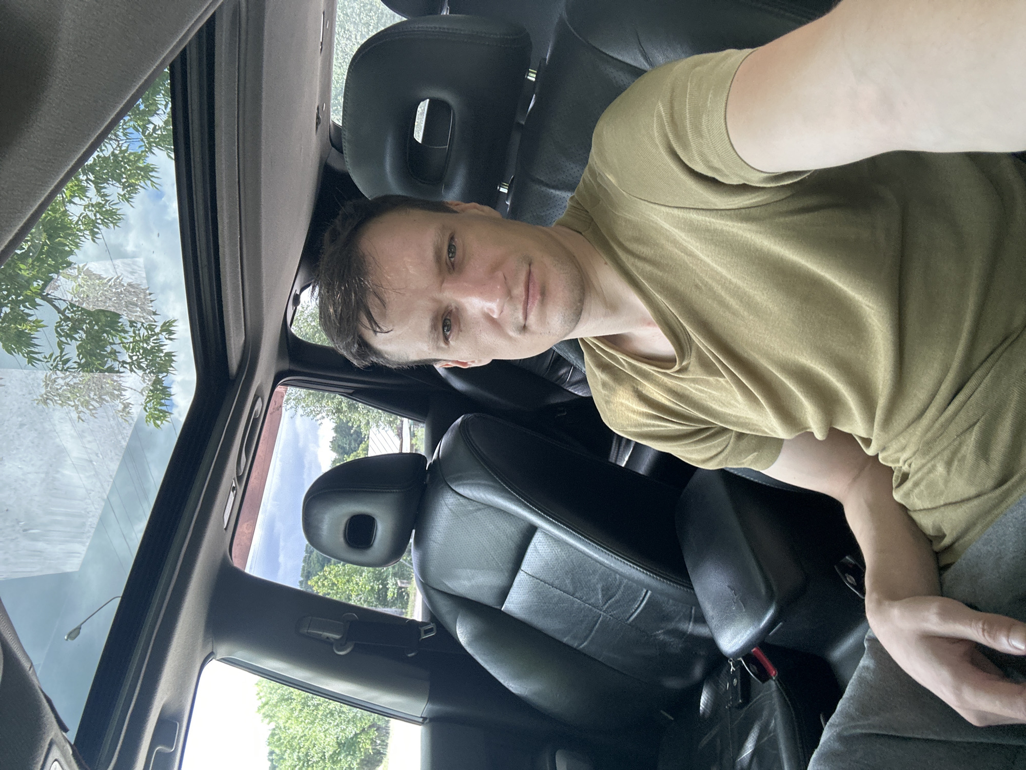 A man is seated in the driver's seat of a vehicle with a sunroof open. He is looking at the camera and appears to be taking a selfie. The interior of the car is visible, including black leather seats and the center console. Outside the car, trees and a cloudy sky are visible. The image was taken in Makiv, Ukraine.