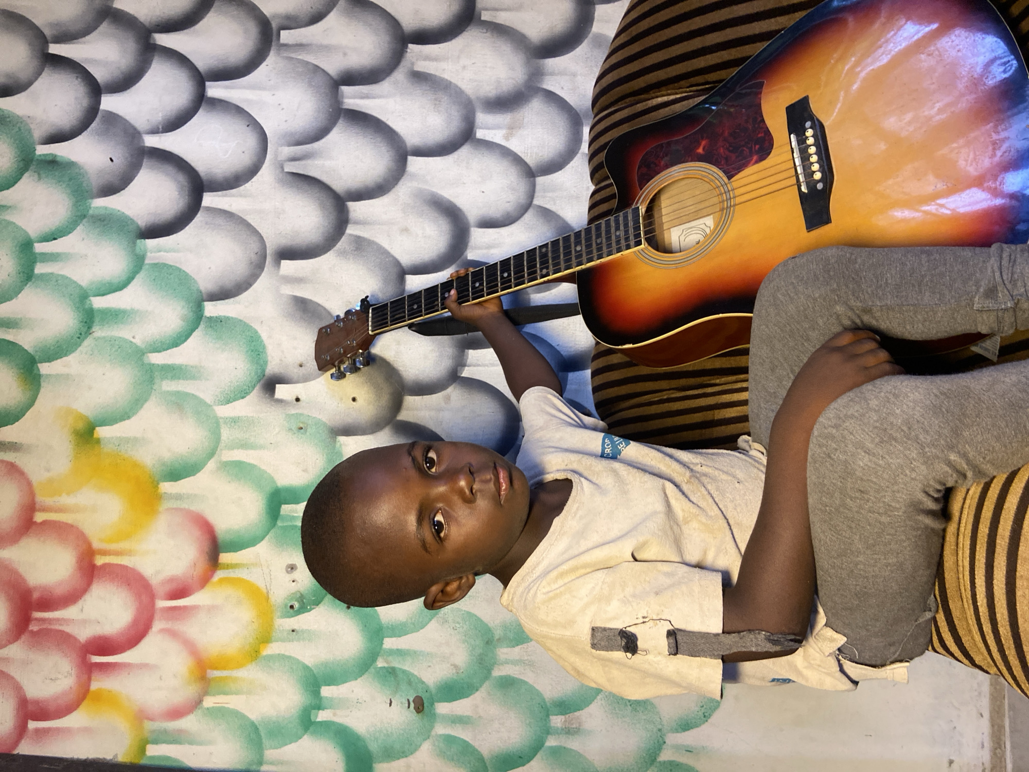 A young boy in Mararaba, Nigeria lies on a striped cushion with an acoustic guitar. He wears a light-colored t-shirt and grey sweatpants. The background features a wall with a scale-like pattern, transitioning from grey at the top to green, yellow, and pink at the bottom. The guitar rests on the cushion beside him.
