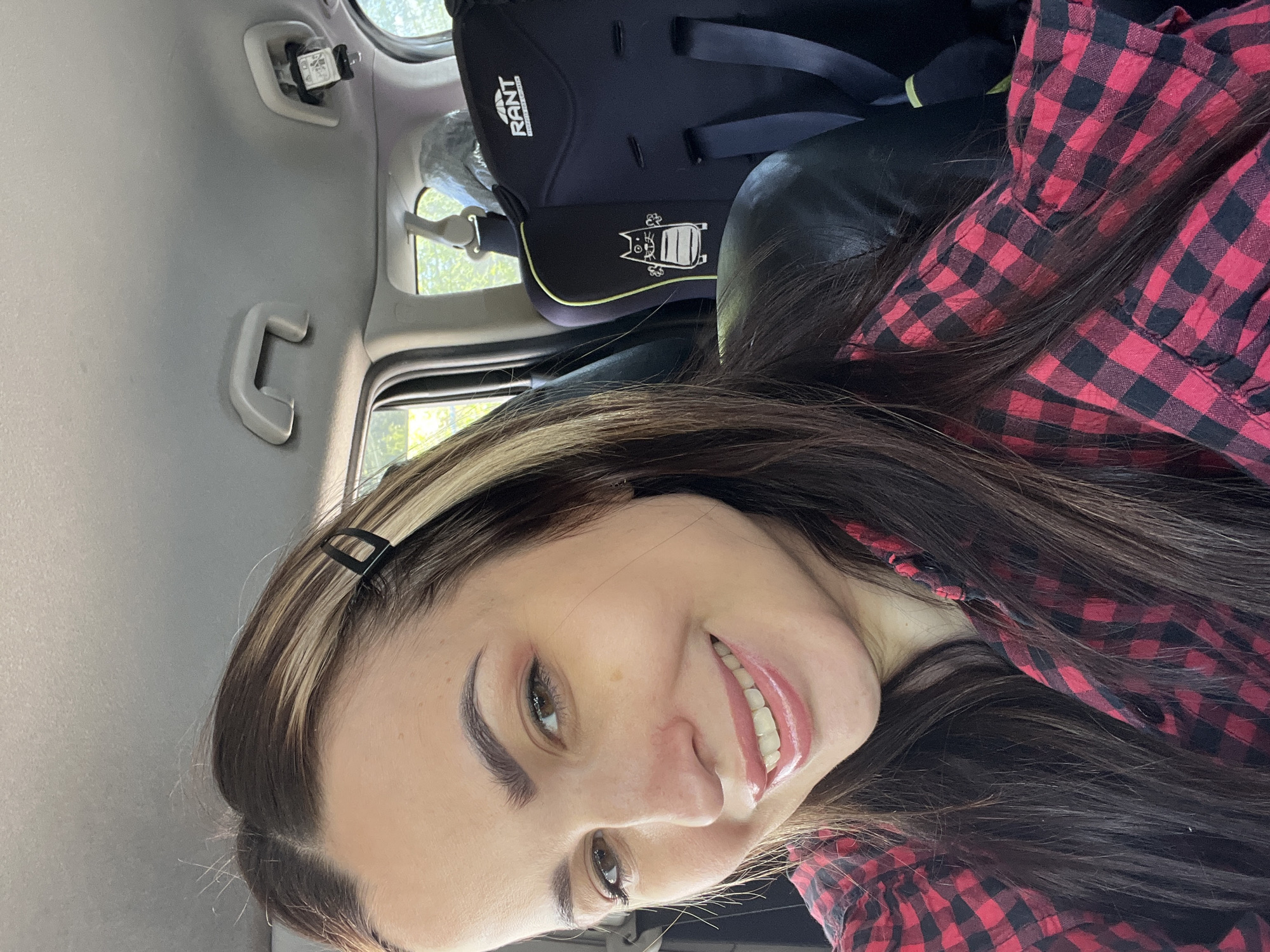 A young woman sits in the back seat of a car. She is smiling and wearing a red and black plaid shirt and a black hair clip. A black child car seat is in the background. The location is Unknown, Unknown.