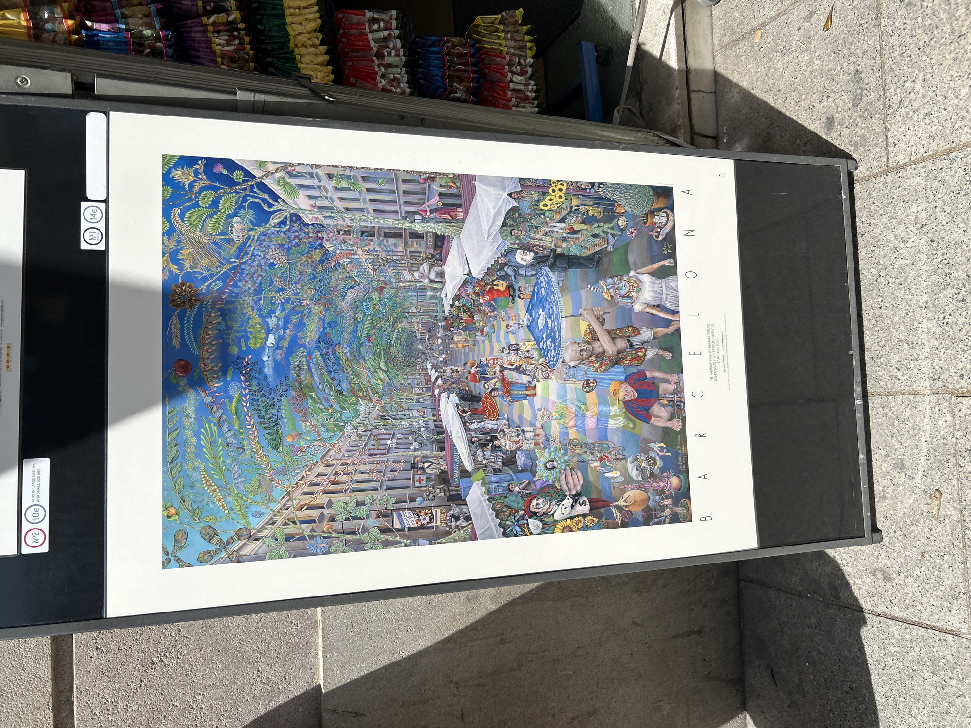 The image depicts a framed poster on display in Barcelona, Spain. The poster showcases a colorful, whimsical painting of Las Ramblas, a famous street in Barcelona, known for its human statues. The painting includes representations of people, buildings, and plants. The poster is displayed on a stand on a sidewalk. The price tags for different sizes of the poster are visible, showing prices of 10 euros for a small size and 14 euros for a large size. A rack above the poster stand holds snacks and sweets.