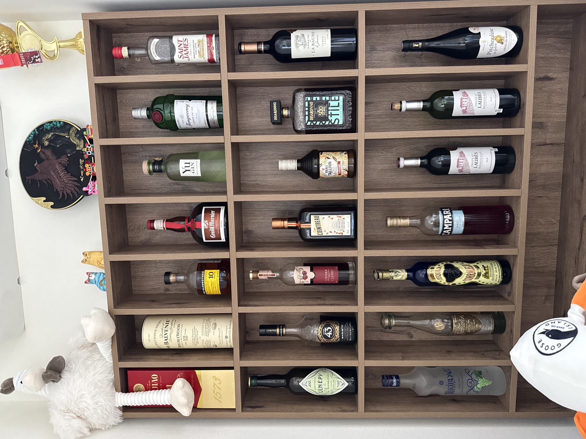 The image shows a wooden shelf unit filled with various bottles, likely alcoholic beverages. The bottles are arranged in a grid-like pattern within the shelf compartments. Labels visible on some of the bottles include "Saint James Rhum Agricole," "Disaronno," "Yu Gin," "Grand Marnier," "The Glenrothes," "Balvenie Doublewood," and "Campari." Several plush toys and other decorative objects are positioned around the shelf unit. The setting is indoors with white walls visible on the sides, suggesting a domestic or commercial space. There is no indication of this being in Sochi, Russia.