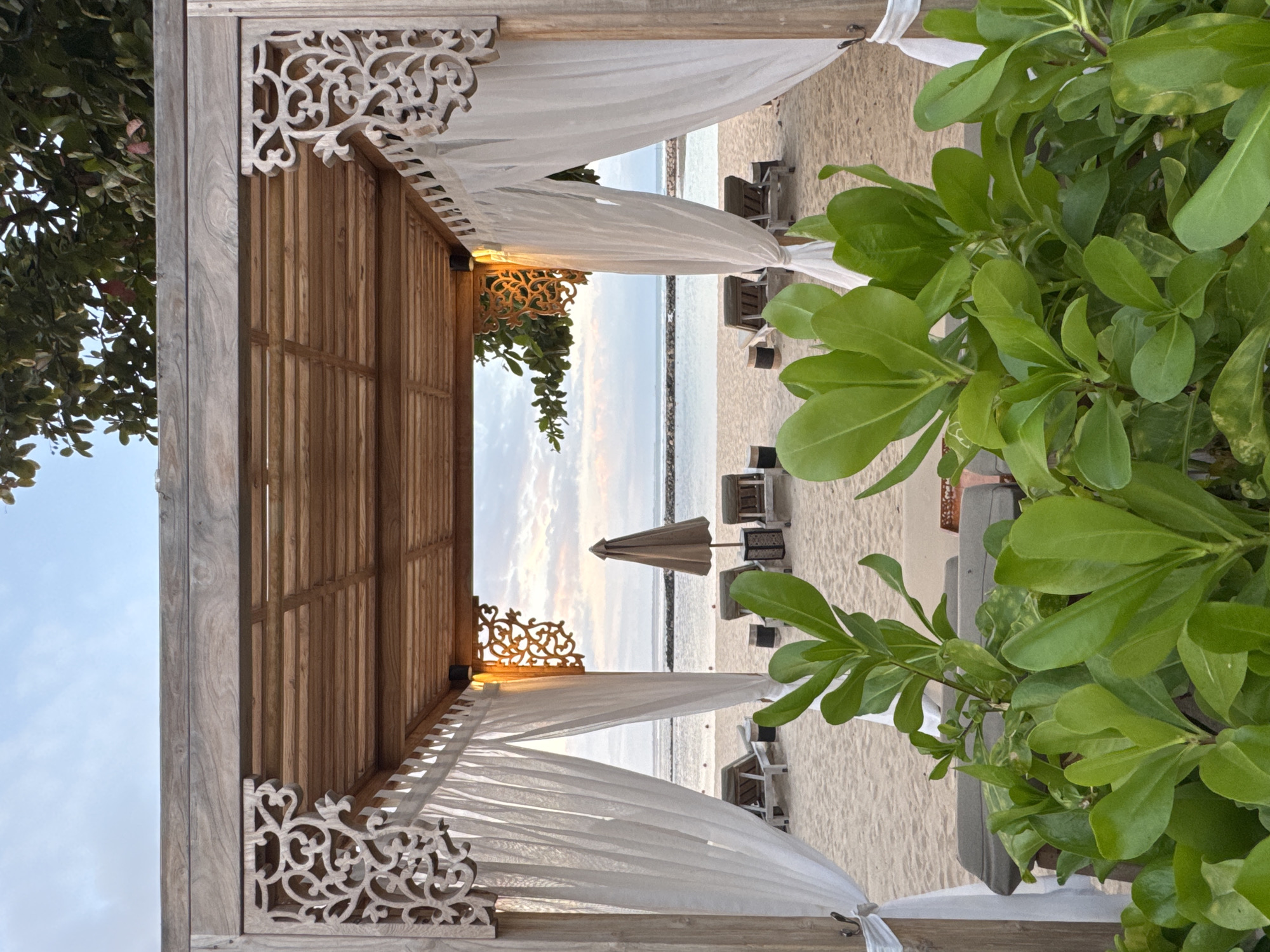 The image shows a view from within a wooden cabana, looking out towards a sandy beach and the ocean. The cabana has a slatted wooden roof and decorative carved wooden panels on the sides. White sheer curtains hang from the sides, framing the view. In front of the cabana, lush green plants are in focus.
Beyond the cabana, the sandy beach is lined with several empty lounge chairs, some with their backs up and some folded. A single large, tan umbrella stands in the middle distance. The ocean is visible in the background, with a breakwater or jetty extending into the water. The sky is overcast with soft clouds, suggesting early morning or late evening light. The scene depicts a tranquil resort setting, likely in Kuta Selatan, Indonesia, a region known for its beaches and tourist accommodations. There are no people visible in the image.