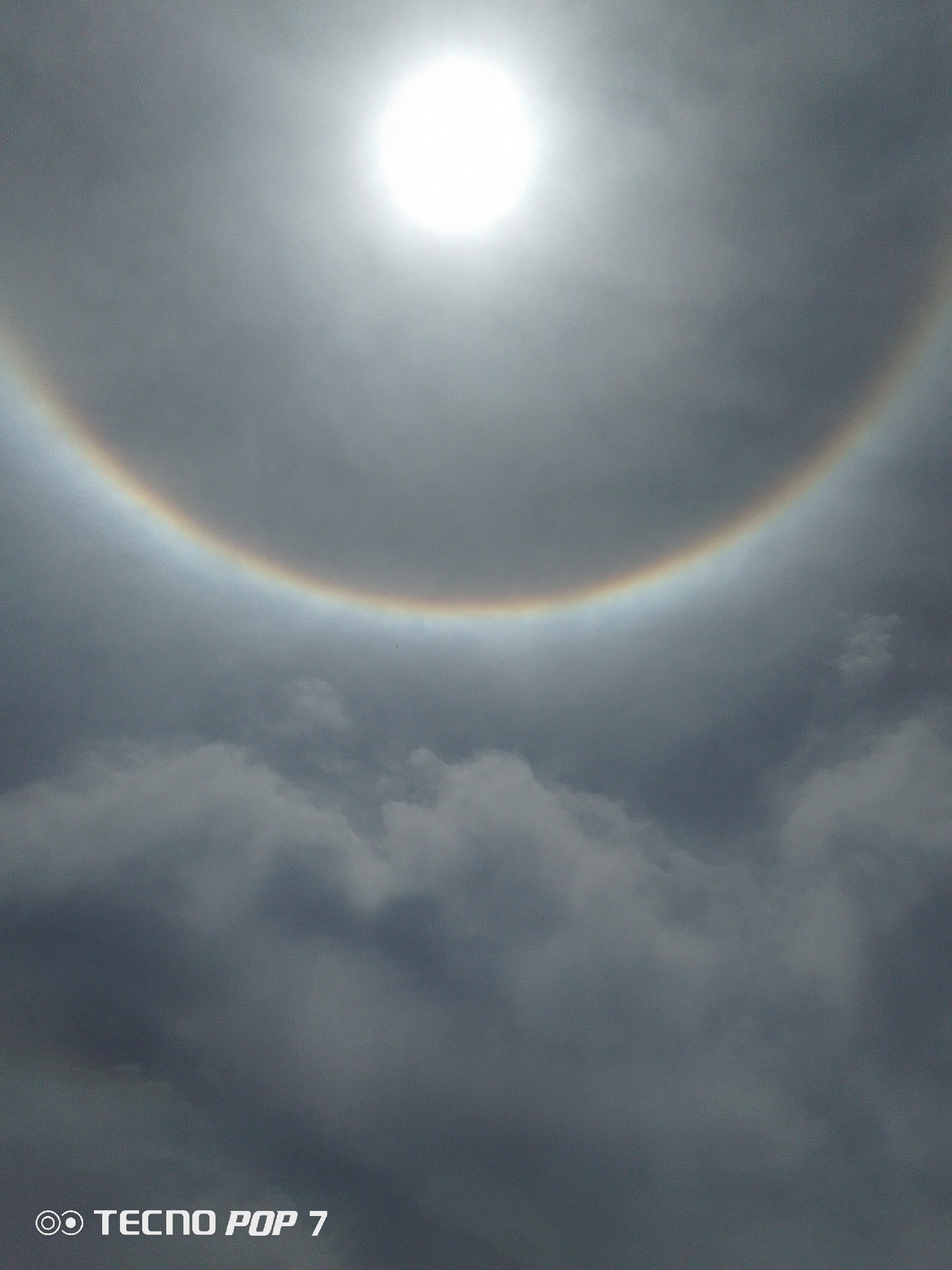 The image depicts a bright sun surrounded by a distinct halo, visible through a sky filled with gray and dark gray clouds. The halo displays a rainbow-like effect. The text "TECNOP POP 7" is visible in the lower-left corner. The location is Pedro González, Venezuela. There are no people or any other objects visible in the image besides the sun, clouds, and halo.