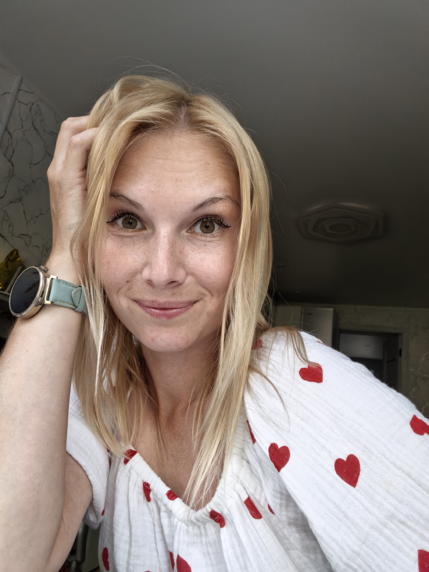 A blonde woman is taking a selfie, resting her hand on her head. She is wearing a white top with red heart patterns and a light blue watch. The background appears to be an indoor setting with a ceiling light fixture visible. The image does not provide any information about the city of Sterlitamak, Russia.
