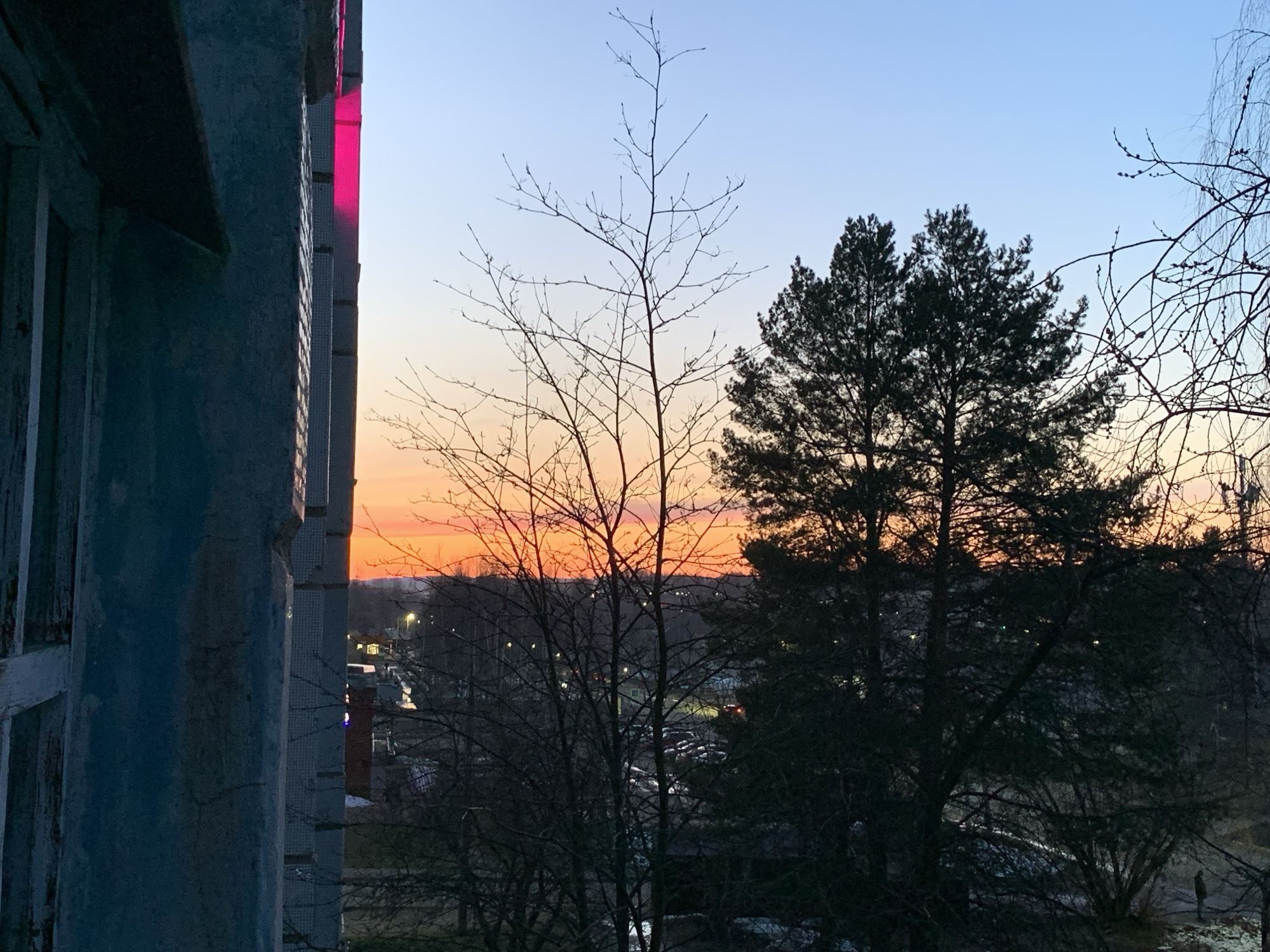 The image depicts a view from a building in Chaikovsky, Russia, at dusk. The sky transitions from orange at the horizon to light blue above. In the foreground, the corner of the building, constructed of grey tiles and old white window frames, is visible to the left. A red light can be seen reflected on the building from the left. Several bare trees and a large evergreen tree dominate the midground, partially obscuring the view of the city. The city consists of residential buildings and lights along the road. In the lower right of the image, a single person appears to be standing and looking towards the city.