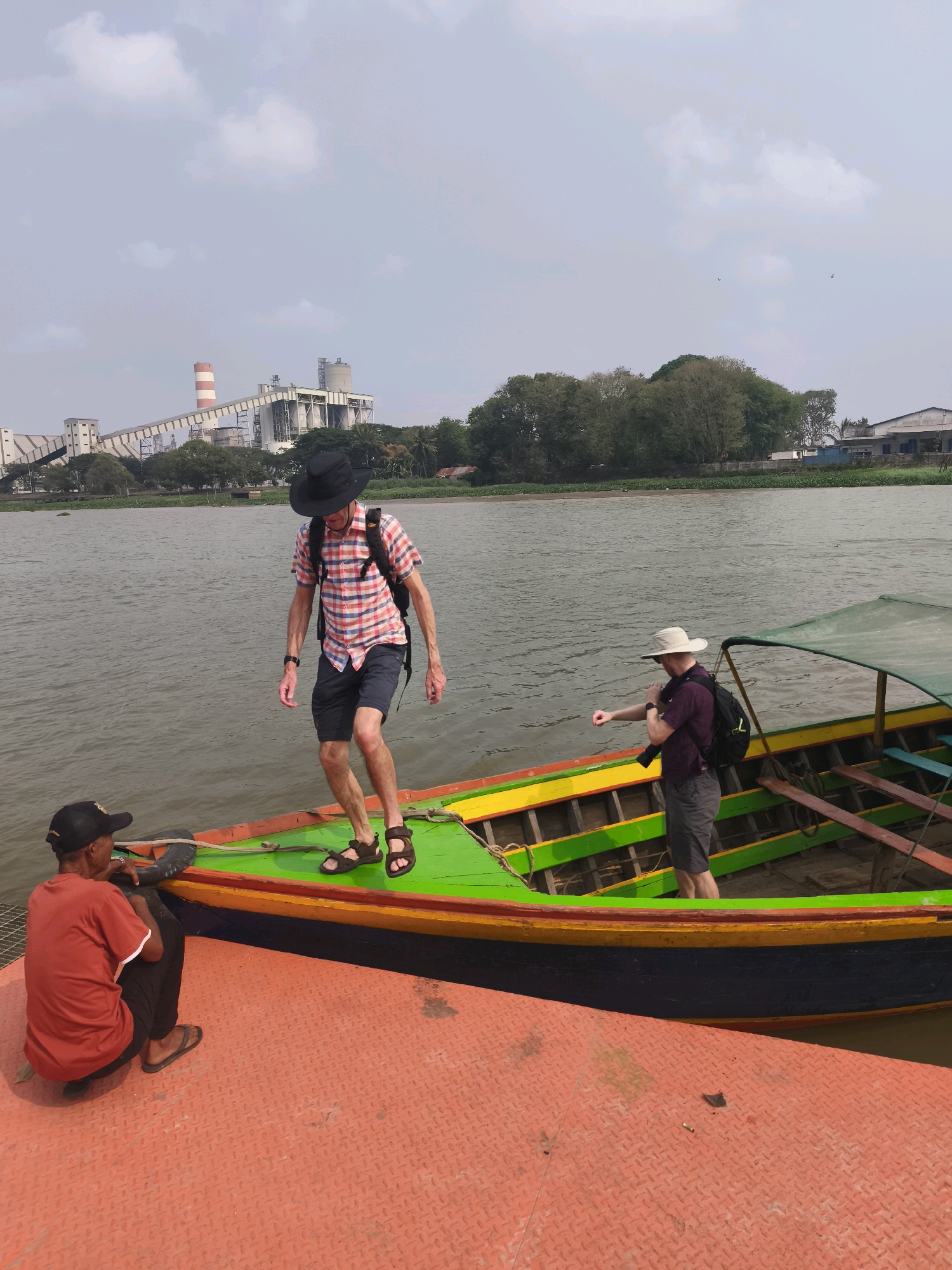 In Palembang, Indonesia, a person wearing a red shirt and black cap is seated on an orange platform next to a brightly colored boat. The boat, painted green, yellow, and red, has a canopy and wooden seats. Two men stand on the boat; one in a plaid shirt and black hat is stepping towards the rear, while another in a purple shirt and white hat is looking away from the camera, possibly at his watch. In the background, a large industrial complex with smokestacks is visible across a body of water, surrounded by trees and buildings. The sky is overcast.