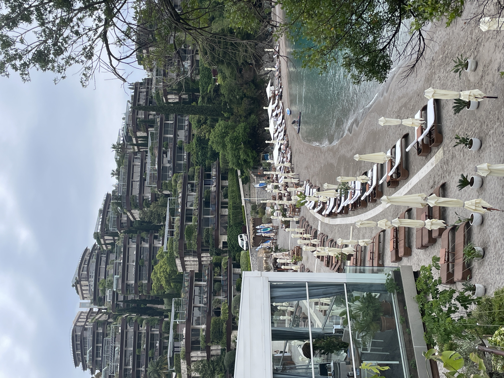 The image shows a coastal resort area in or near Budva, Montenegro. A multi-level building complex with verdant vegetation covering the balconies and roofs is on the left. A sandy beach curves along the coastline, lined with rows of beach chairs and umbrellas. The sea is calm with gentle waves. The lower right of the image includes the corner of a glass-enclosed structure, possibly a restaurant or lounge area. People are seen on the beach, some near the water's edge.
