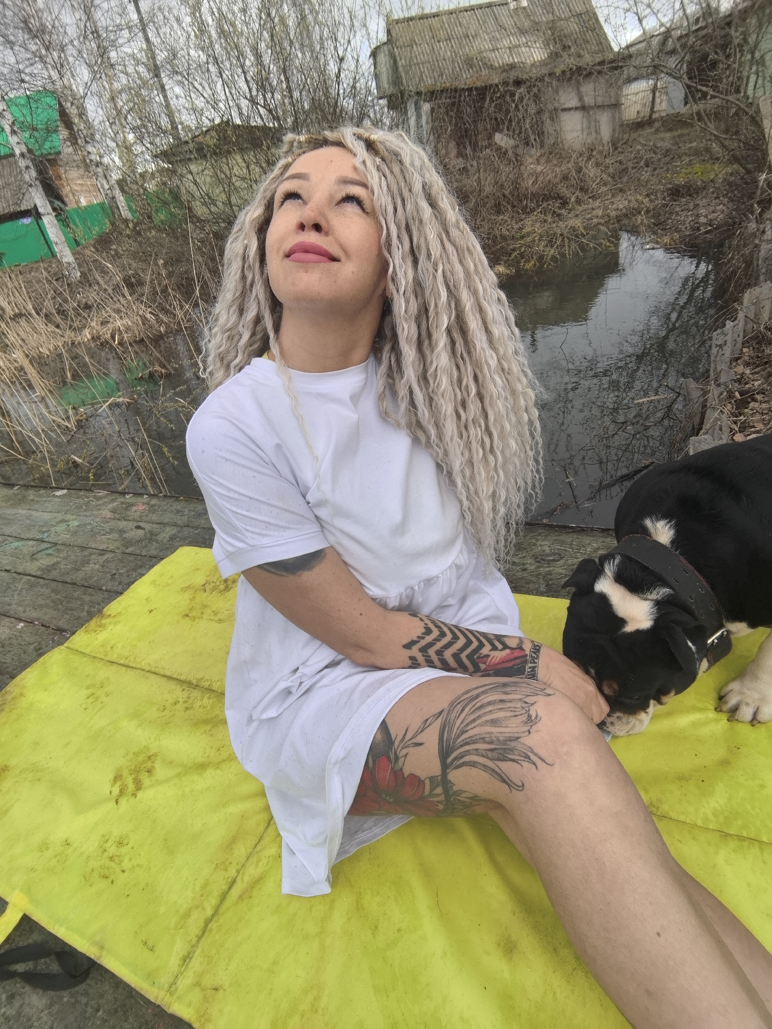 A woman with long, light blonde dreadlocks is seated on a bright yellow mat on what appears to be a wooden platform or dock. She is wearing a simple white dress or oversized t-shirt. Her head is tilted slightly back as she looks upwards with a contemplative expression and a subtle smile. She has visible tattoos on her left arm, including a geometric pattern and the text "TWIN PEAKS," and a large floral or leafy design on her upper left leg.
Beside her, to her right, a black and white dog, resembling a bulldog, is present. The dog's head is lowered towards the yellow mat, appearing to sniff or interact with something on its surface. The yellow mat shows signs of wear, with visible dirt or stains.
The setting is outdoors, next to a body of still water that reflects the overcast sky. The background is rural and somewhat unkempt, featuring numerous bare trees and dry vegetation, suggesting a late winter or early spring season. Beyond the trees, several old, rustic buildings or shacks with weathered roofs are visible, adding to the rural aesthetic. The sky is uniformly grey and overcast, indicating daytime. The overall scene depicts a quiet moment between a woman and her dog in a natural, rustic environment in Noviy, Russia.