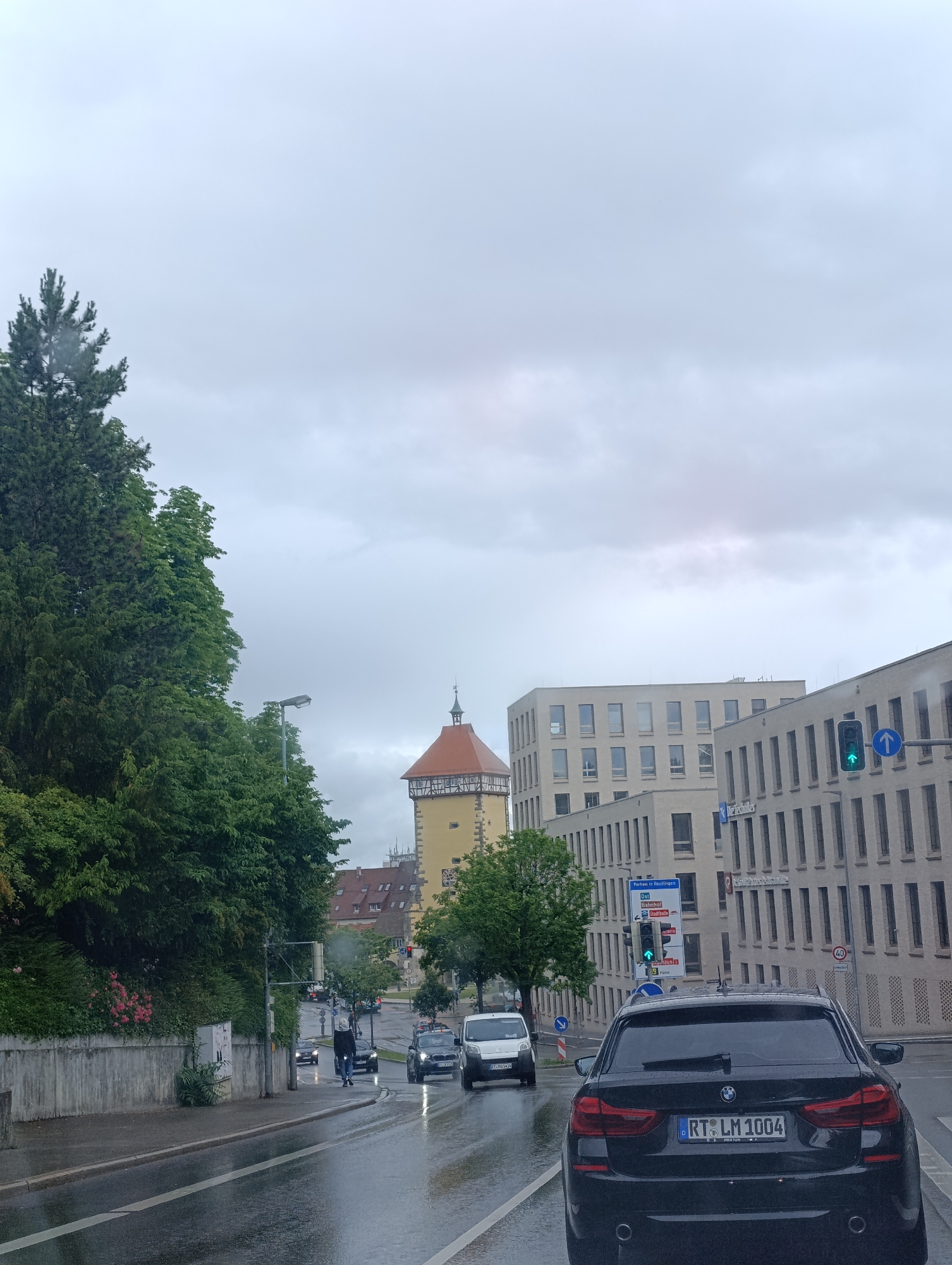 The image depicts a street scene in Reutlingen, Germany, on a cloudy day, possibly during or after rainfall, as the road surface is wet. In the foreground, a black BMW station wagon with license plate "RT LM 1004" is the primary subject, facing away from the camera. To the left of the BMW, a white van is visible. The road features distinct lane markings.
Dominating the left side of the image is a dense cluster of lush green trees, with a concrete wall bordering the sidewalk. Further down the street, a yellow tower with a red roof, characteristic of a historical building, stands against the sky. Modern, light-colored buildings with numerous windows are prominent on the right side of the street. A traffic light displaying green and a blue directional sign indicating "Parken in Reutlingen" with various destinations like "Ost," "Bahnhof," and "Eisdielen" are also visible.
Several other vehicles, including cars and a person walking in the street, are present further down the road. The sky is overcast, contributing to the subdued lighting of the scene.